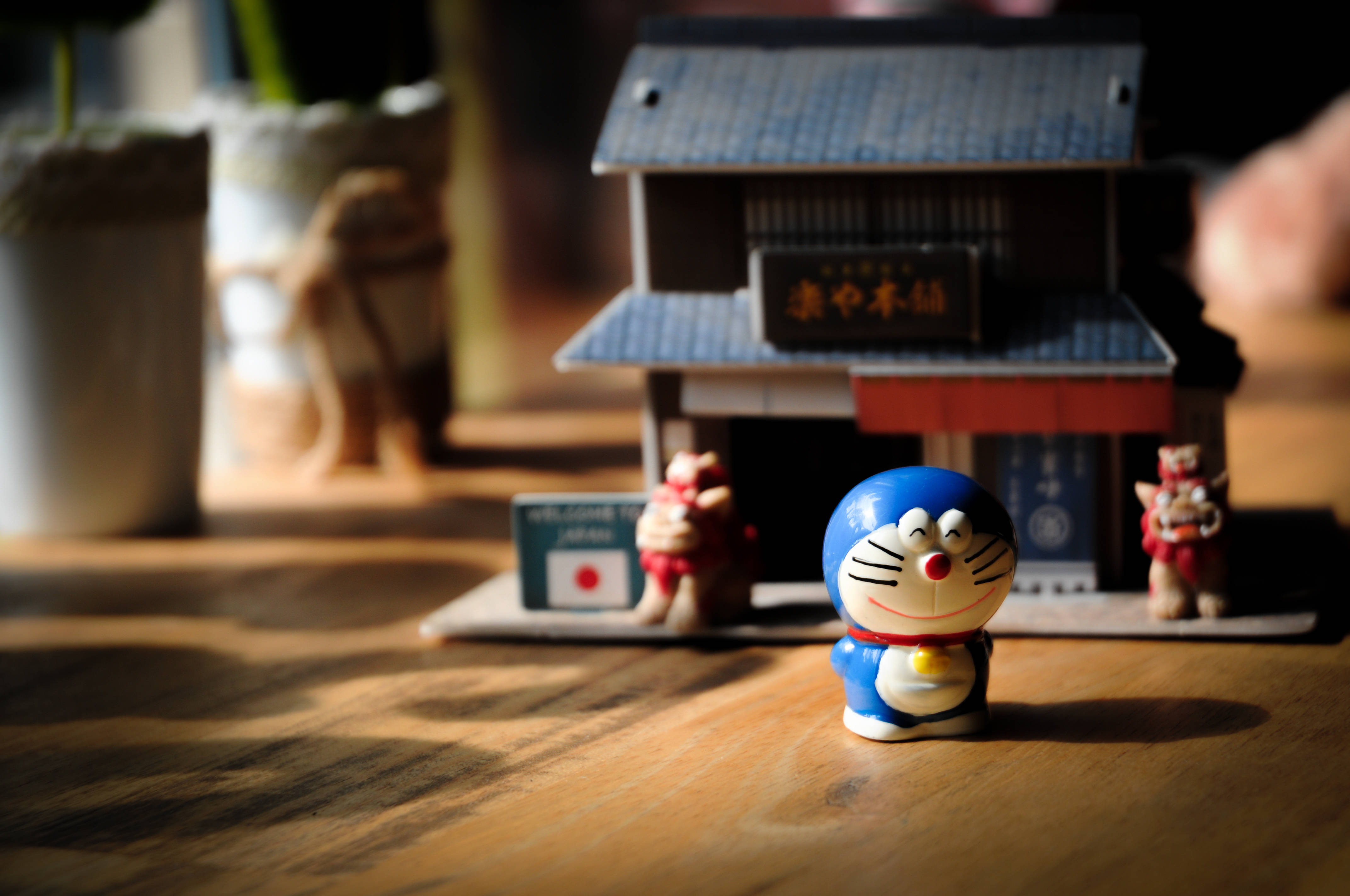 Doraemon 桌布 壁紙 — 一個藍色多啦A夢公仔坐在木桌上，旁邊有一個帶有紅色燈籠和歡迎牌的微型日本風格房屋。
