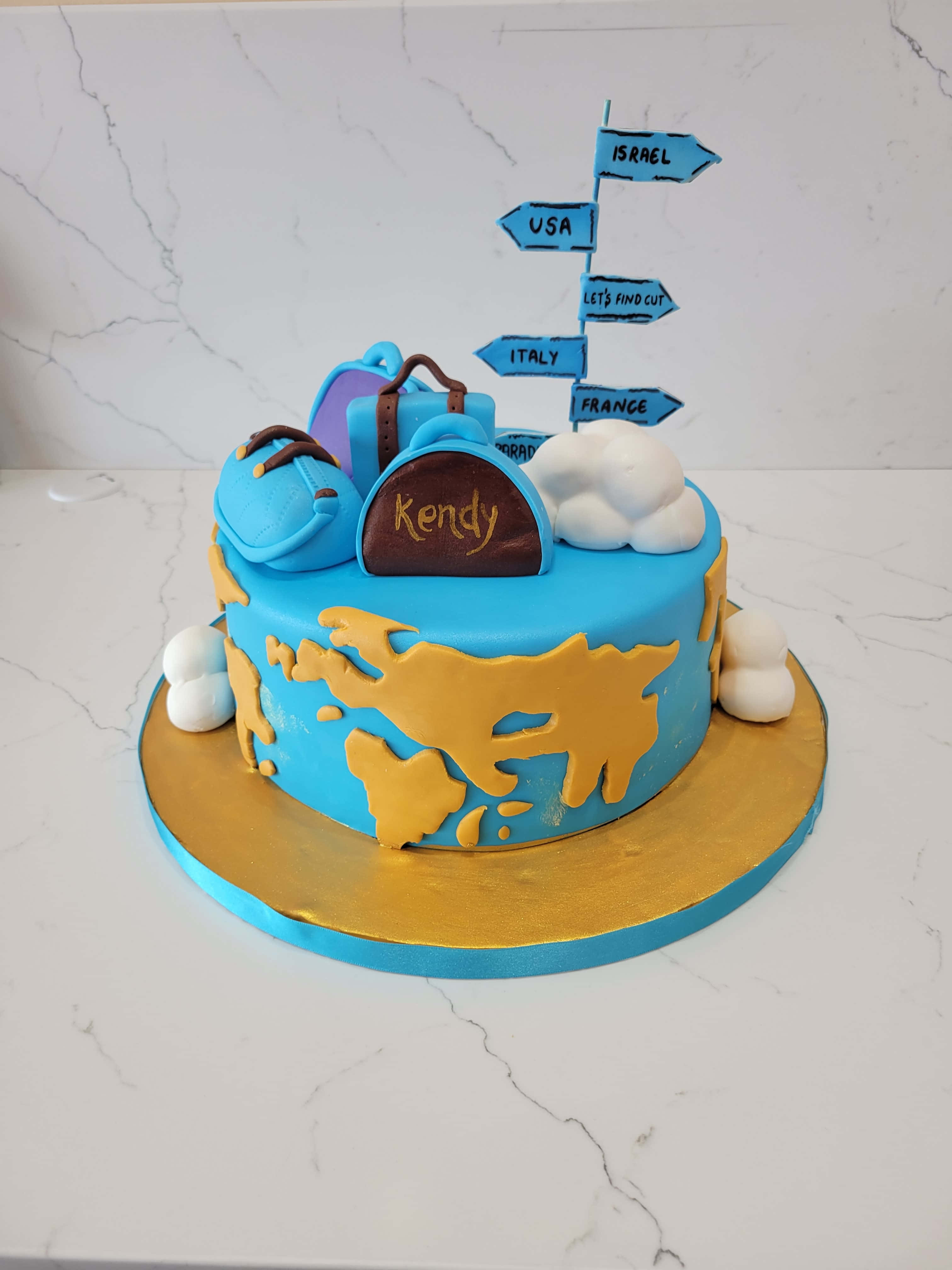 Cake Fondant 桌布 壁紙 — 一個藍色翻糖蛋糕，裝飾有金色輪廓的世界地圖和旅行主題裝飾，包括行李箱、雲朵和指向國家的方向標誌。