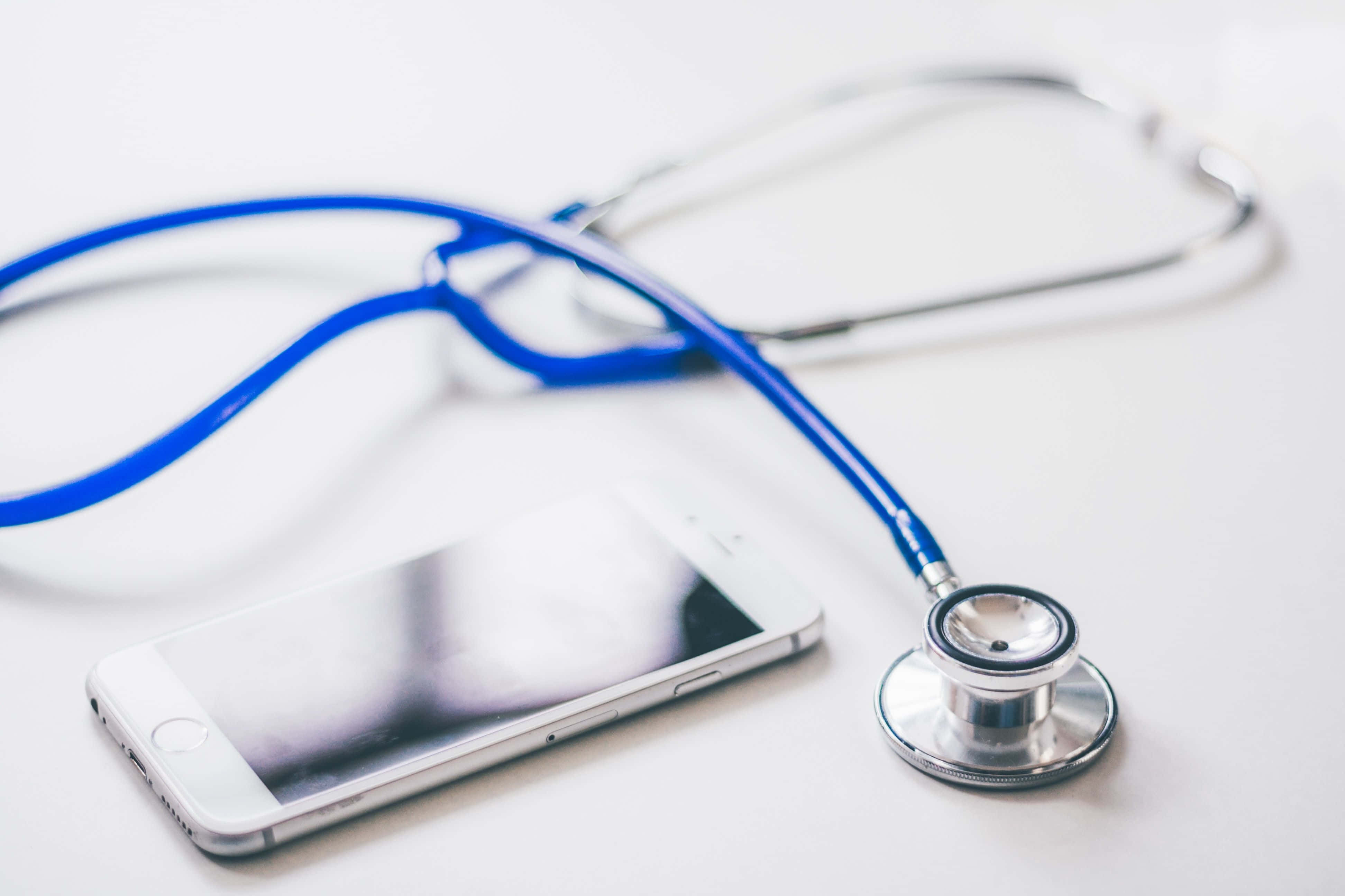 Stethoscope 桌布 壁紙 — 一個藍色聽診器放在一部白色智能手機旁邊，置於潔白表面上，帶有柔和陰影。