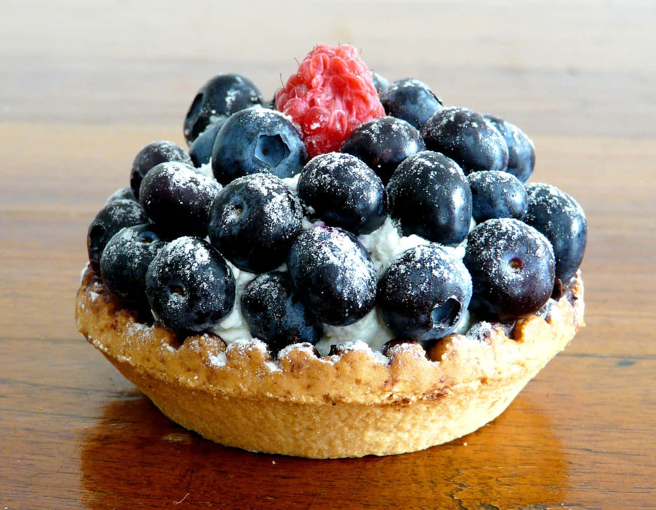 Blueberries Tart 桌布 壁紙 — 一個藍莓撻的近距離特寫，上面撒有糖粉和單粒覆盆子，放在木桌上。
