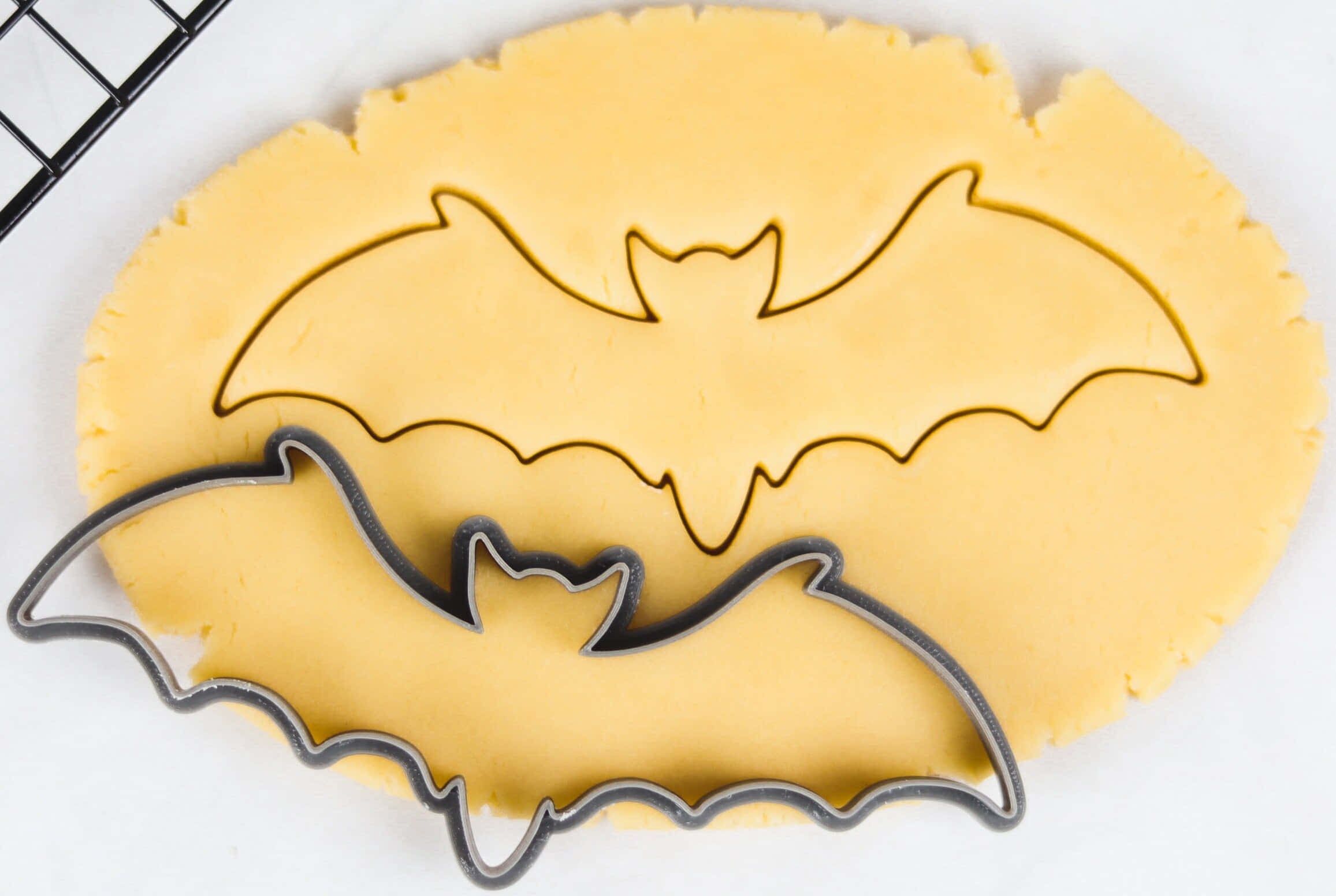 Halloween Cookie Cutters 桌布 壁紙 — 一個蝙蝠形狀的餅乾模具壓在淡黃色的麵團上，白色表面可見冷卻架邊緣。