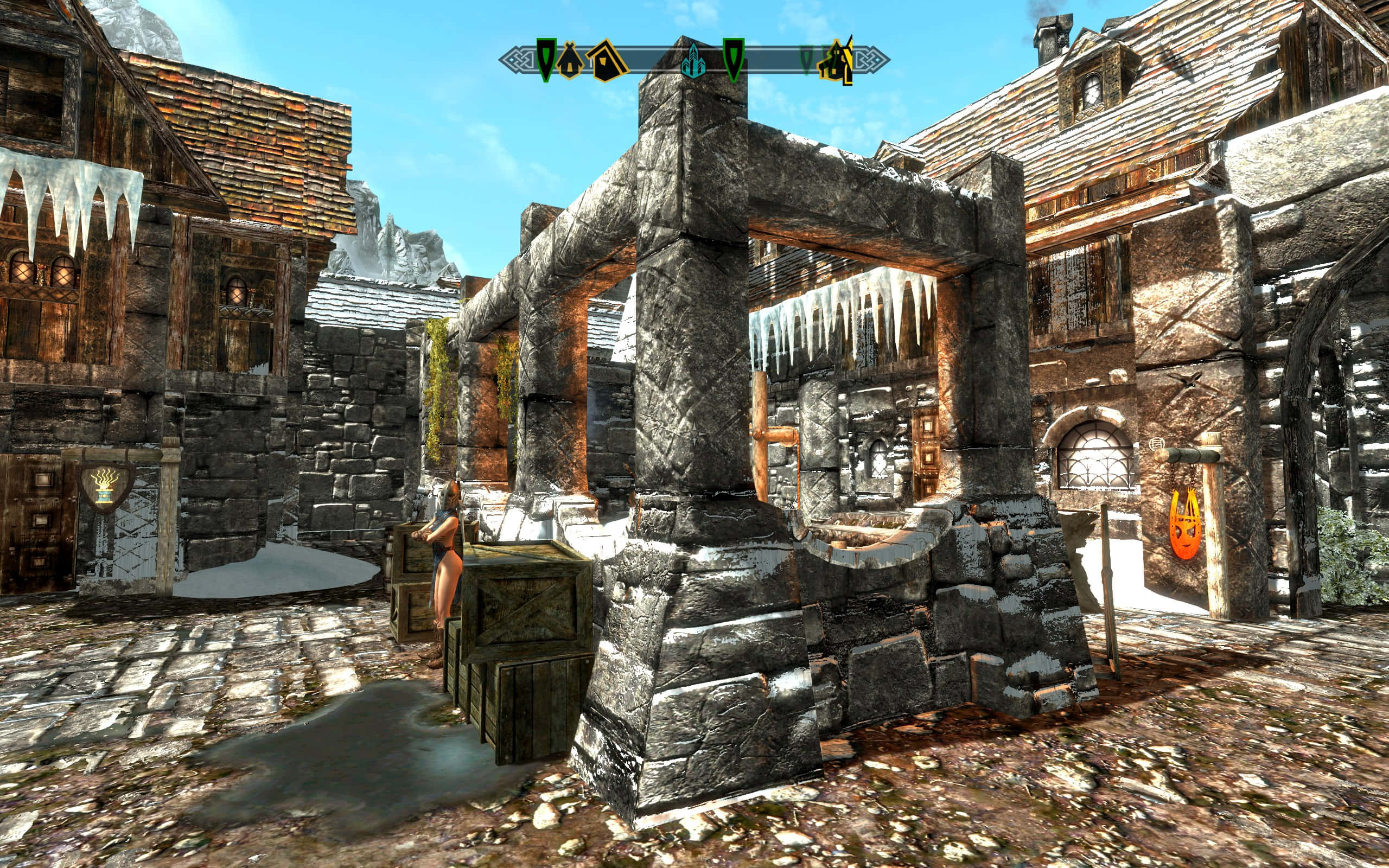 Skyrim Windhelm 桌布 壁紙 — 一個被雪覆蓋的中世紀村莊廣場，有石頭建築、屋頂上垂下的冰柱和鵝卵石街道，在清澈的藍天下。