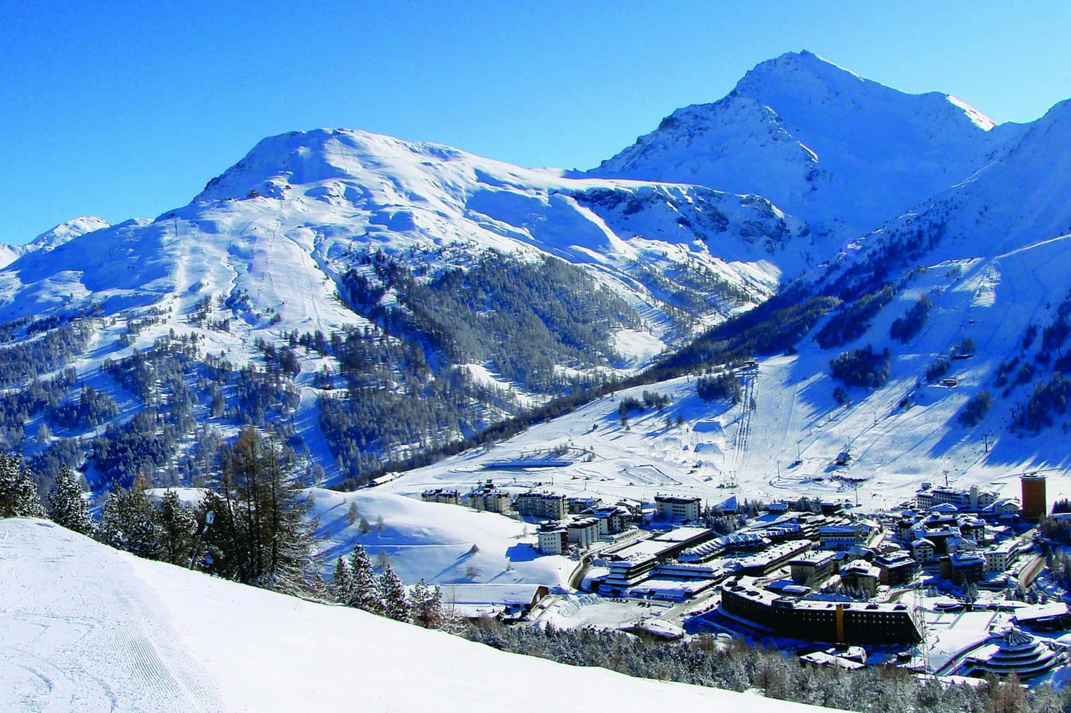 Ski Resort 桌布 壁紙 — 一個被雪覆蓋的山村，坐落在山谷中，在清澈的藍天下，可以看到滑雪道。