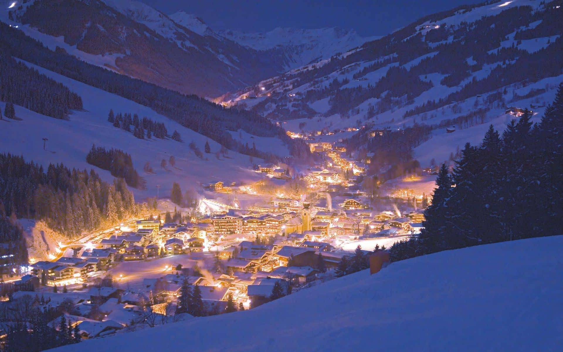 Snowy Village 桌布 壁紙 — 一個被雪覆蓋的山村在夜晚溫暖地發光，坐落於被黑暗松林和雪點綴的山峰環繞的山谷中。