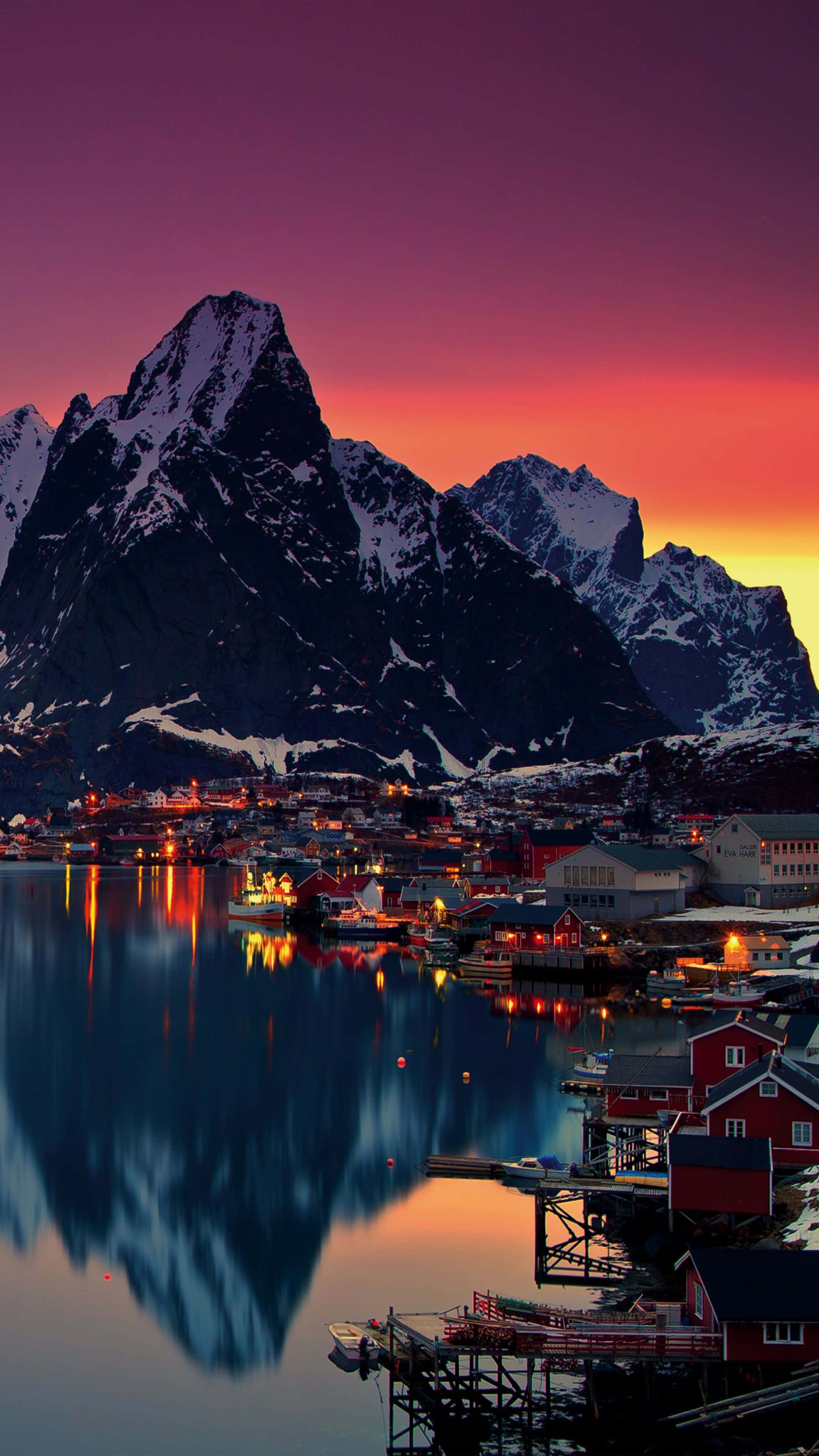 Norway 桌布 壁紙 — 一個被雪覆蓋的山村在日落時分倒映在平靜的水面上，紅色房屋散發出溫暖的燈光。