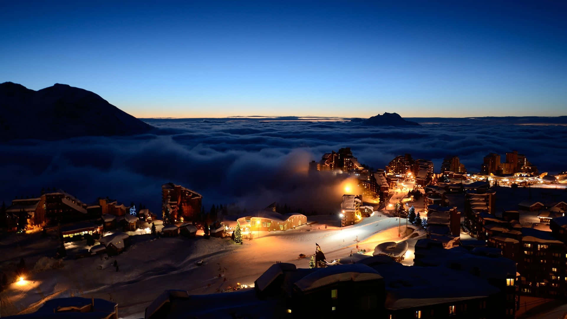 Ski Resort 桌布 壁紙 — 一個被雪覆蓋的山村在暮色中溫暖地發光，位於雲海之上，黑暗的山峰在深藍色的天空襯托下形成剪影。