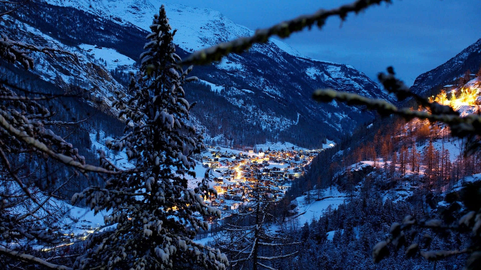Snowy Village 桌布 壁紙 — 一個被雪覆蓋的山村在黃昏時分溫暖地發光，被結霜的松樹枝條框住。