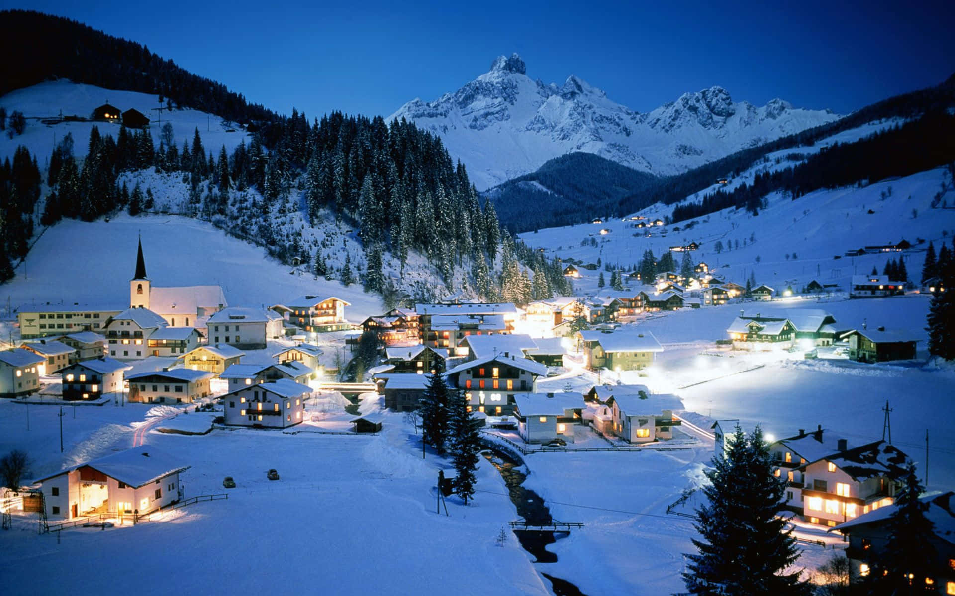Snowy Village 桌布 壁紙 — 一個被雪覆蓋的山村在黃昏時分溫暖地發光，窗戶和教堂尖頂的燈光映襯著高聳的山峰。