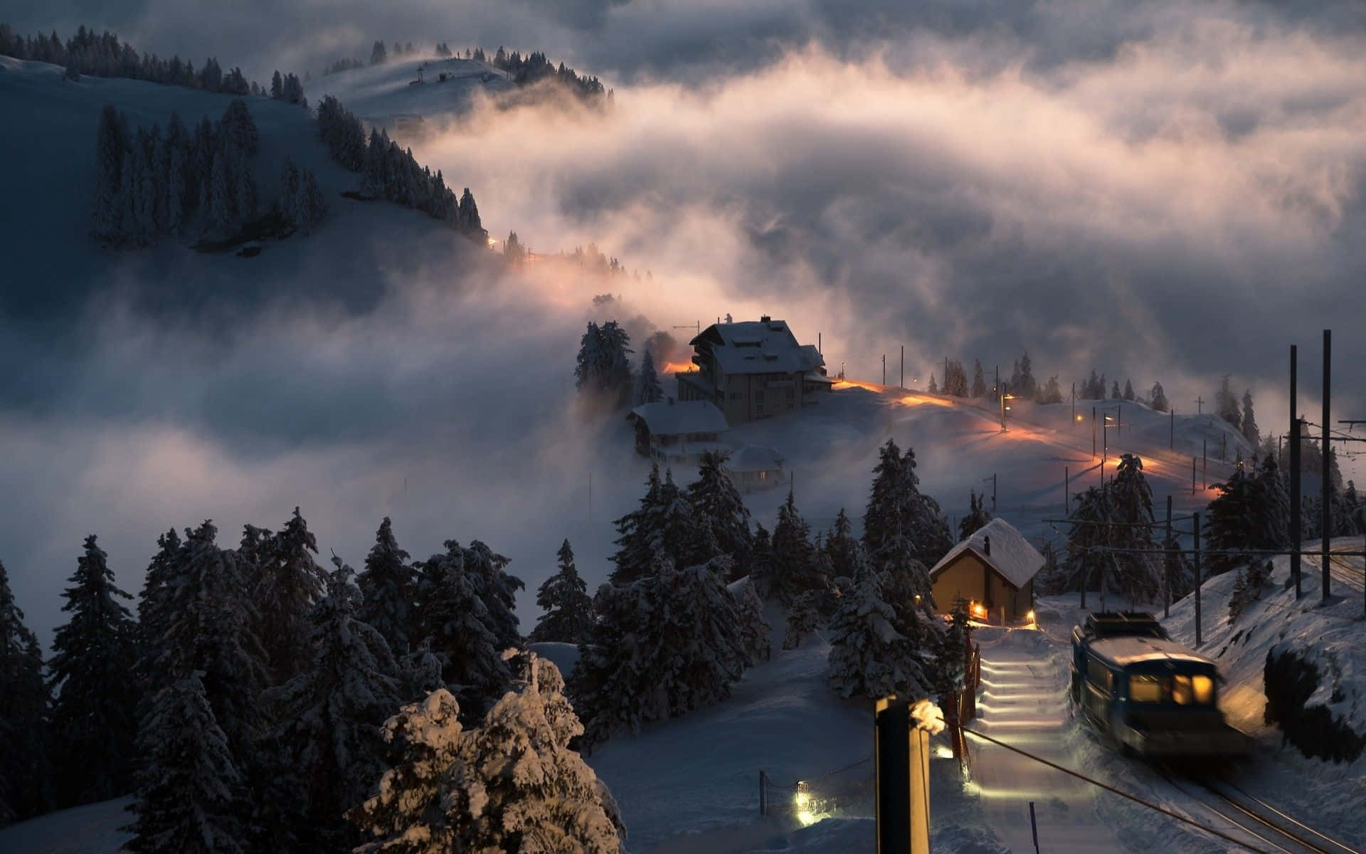 Snowy Village 桌布 壁紙 — 一個被雪覆蓋的山間車站溫暖地發光，一列火車穿過霧氣瀰漫的黎明雲層。