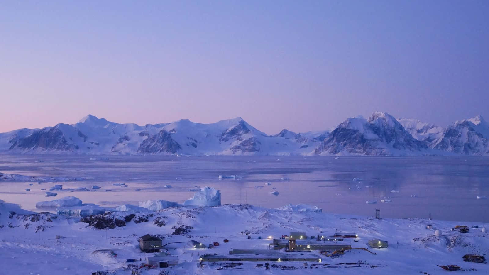 Antarctica 桌布 壁紙 — 一個被雪覆蓋的研究站在暮色中，以溫暖的燈光映照著冰封的山脈和漂浮的冰山。