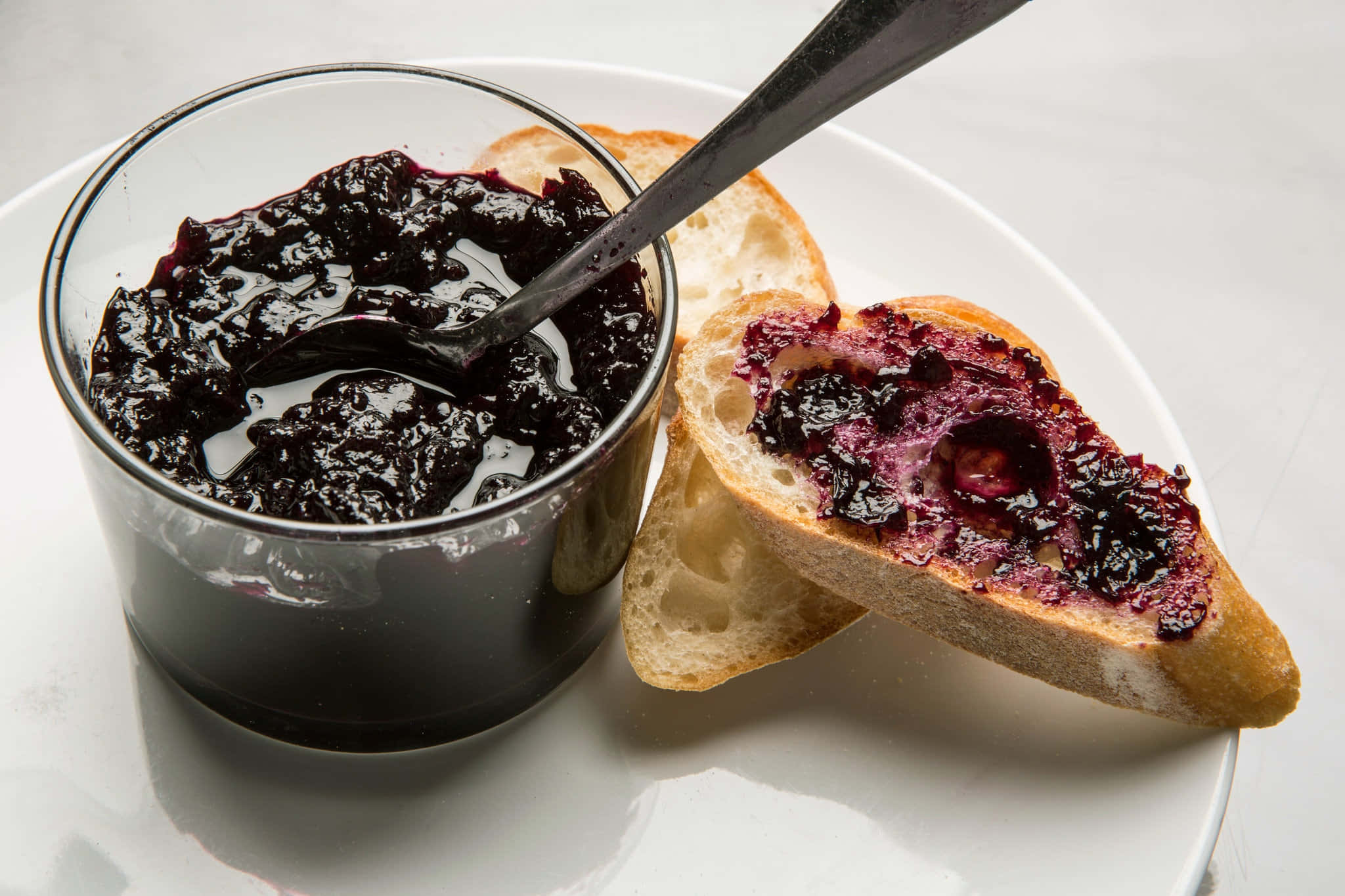 Blueberry Jam 桌布 壁紙 — 一個裝有深紫色莓果醬的玻璃罐和勺子放在白色碟子上，旁邊是一片塗了果醬的脆皮麵包。