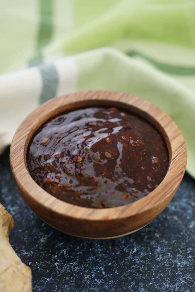 Szechuan Sauce 桌布 壁紙 — 一個裝有濃郁深色醬汁、帶有紅色斑點的醬汁放在木碗裡，置於深色斑點表面上，後面有一塊綠色條紋布處於柔焦狀態。