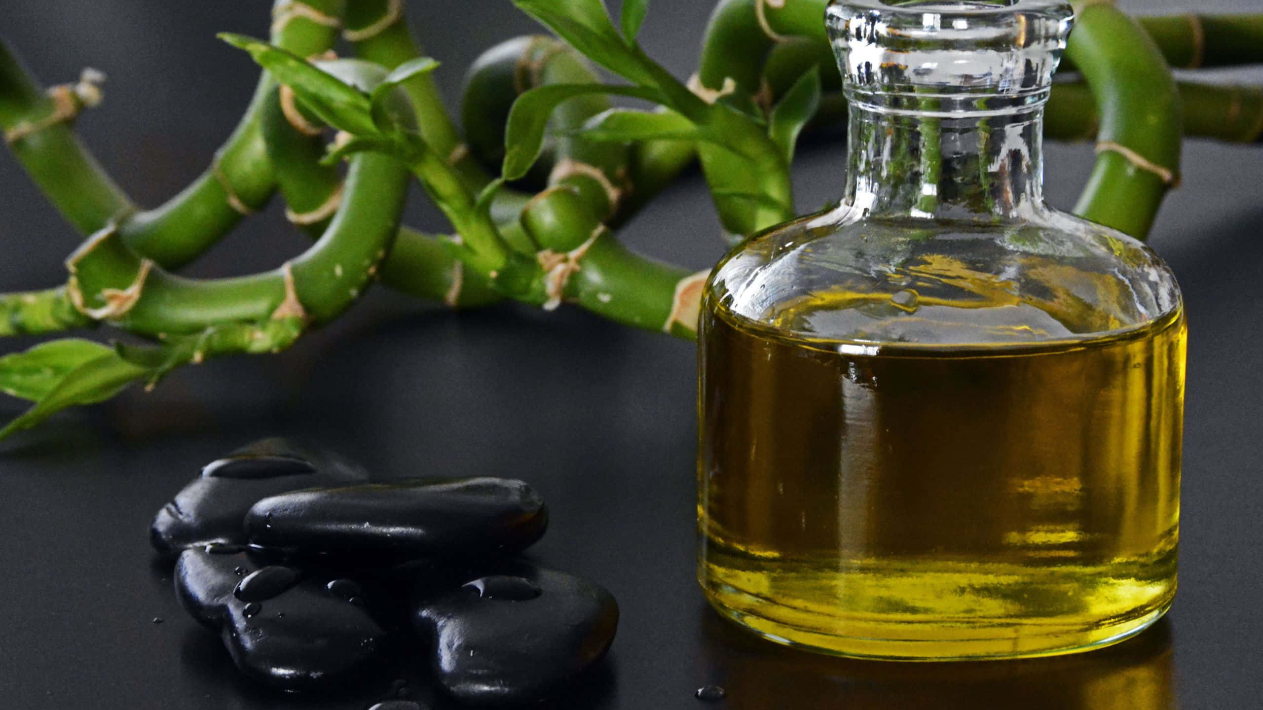 Oil 桌布 壁紙 — 一個裝有金色油的玻璃瓶放在深色表面上，旁邊有光滑的黑色石頭和一棵綠色竹植物。