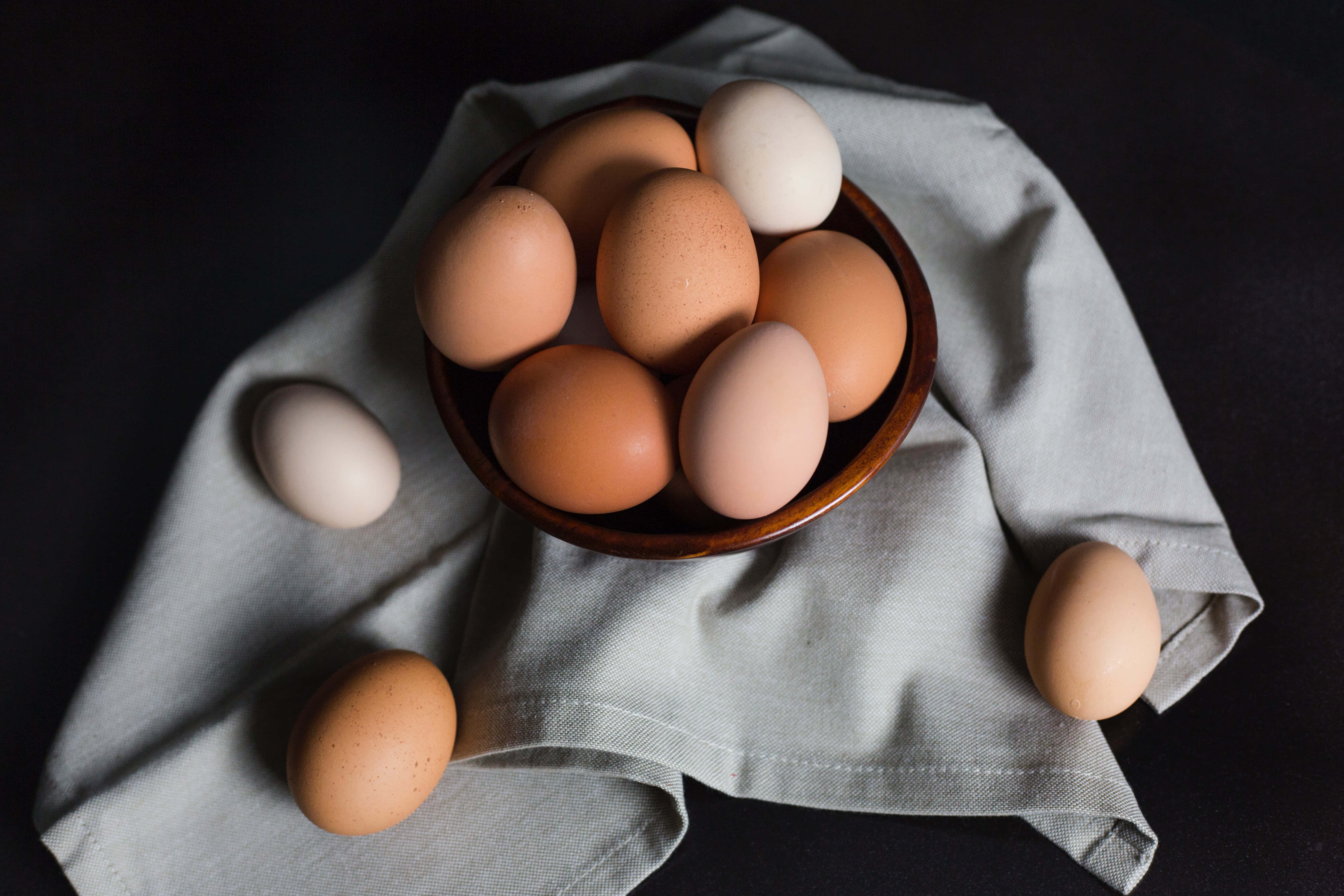 Brown Eggs 桌布 壁紙 — 一個裝滿各種棕色和白色雞蛋的木碗放在淺灰色布上，背景為深色。