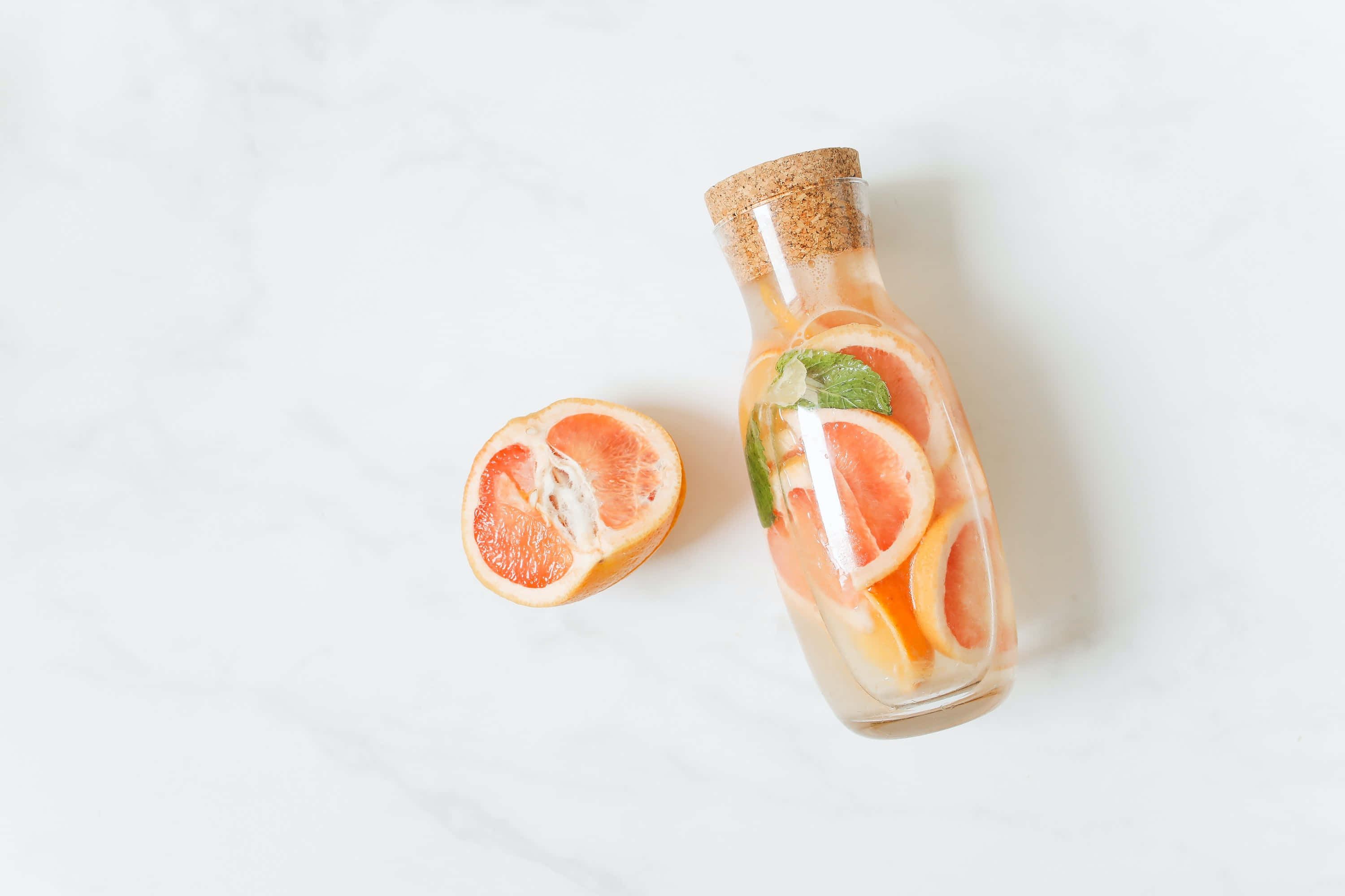 Summer Drinks 桌布 壁紙 — 一個裝滿柑橘片和薄荷的玻璃瓶放在白色大理石表面上，旁邊是一個切半的西柚。