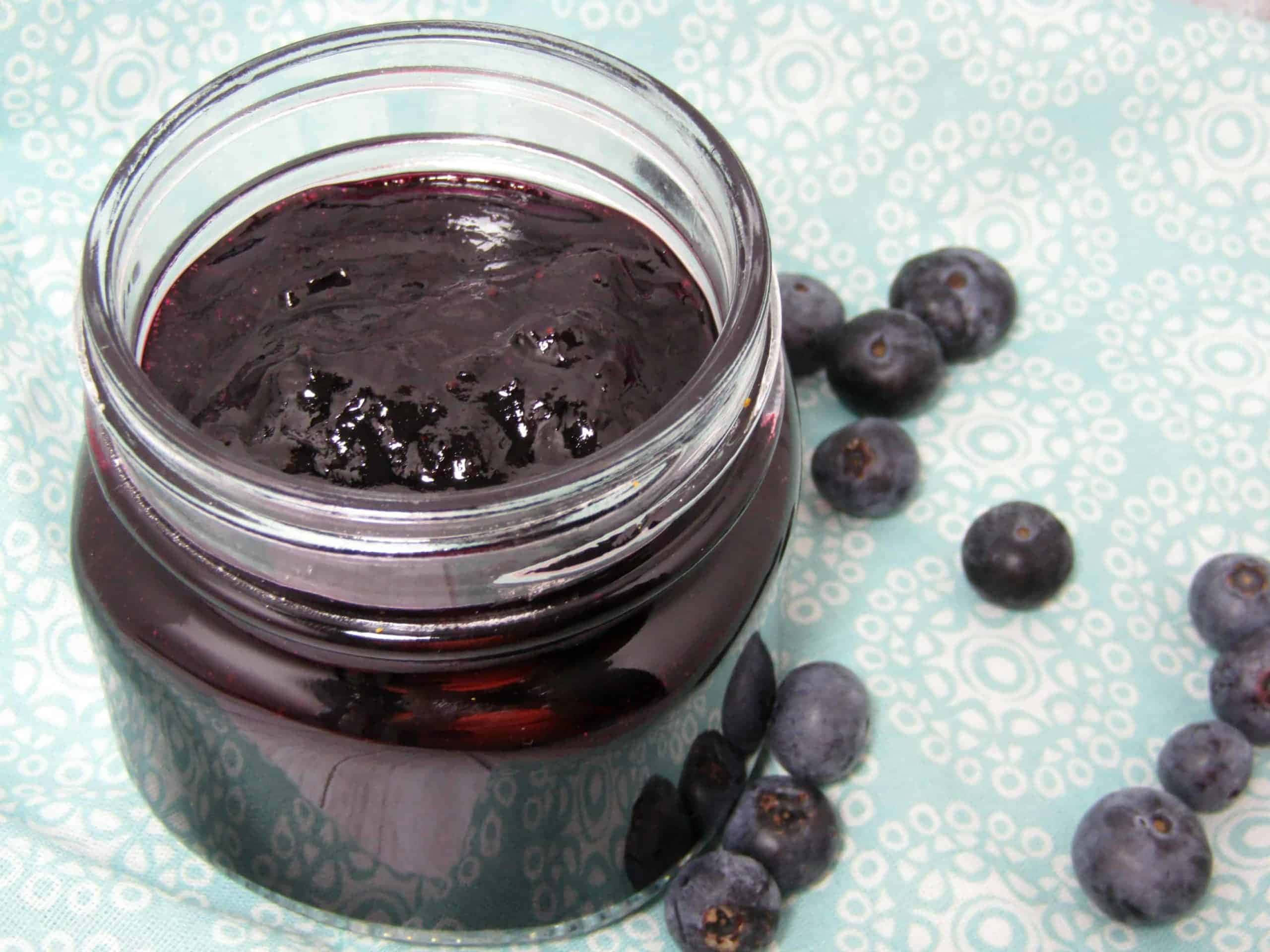 Blueberry Jam 桌布 壁紙 — 一個裝滿濃郁深色藍莓醬的玻璃罐，旁邊放著新鮮藍莓，置於淺藍色圖案布上。