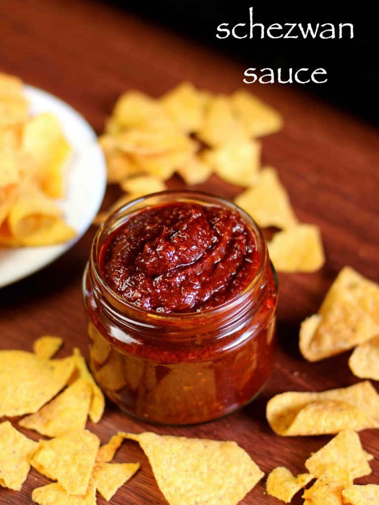 Szechuan Sauce 桌布 壁紙 — 一個裝滿濃郁紅色四川醬的玻璃罐，被金色玉米片包圍，放在木質表面上。