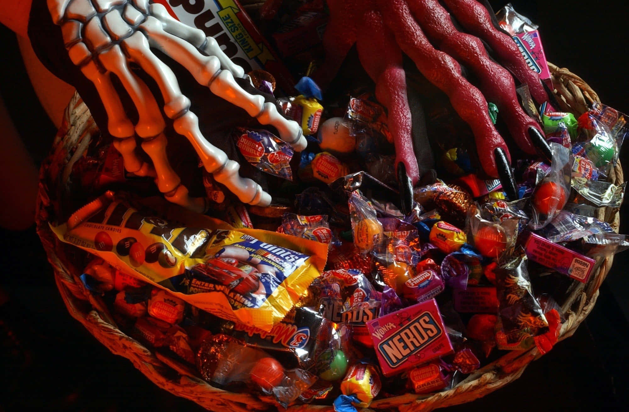 Halloween Treats 桌布 壁紙 — 一個裝滿色彩繽紛萬聖節糖果的籃子，被一隻白色骷髏手和一隻黑色指甲的紅色橡膠手抓住。