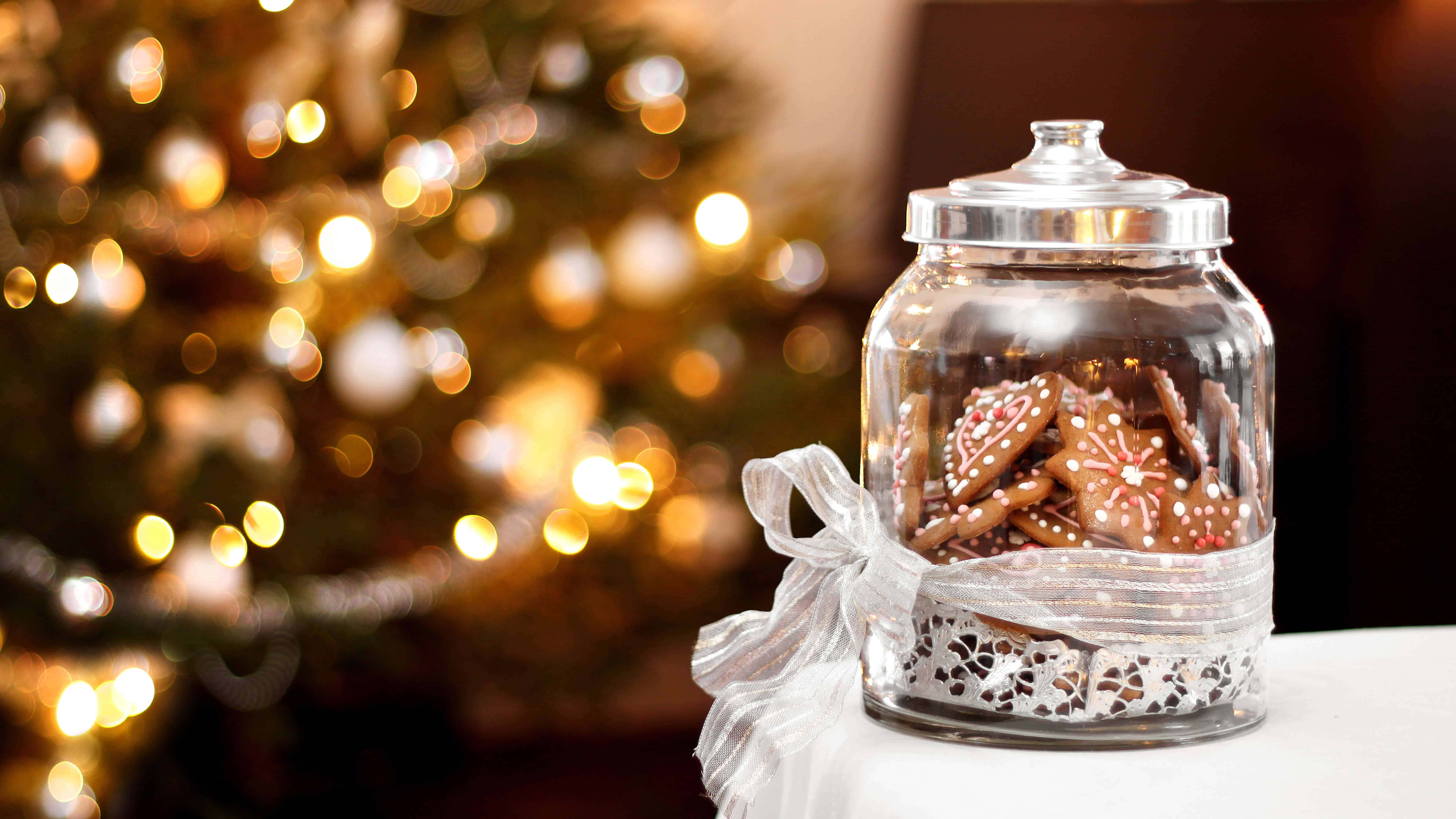 Gingerbread 桌布 壁紙 — 一個裝滿裝飾薑餅曲奇的玻璃罐放在白色表面上，背景是模糊的金色聖誕樹。