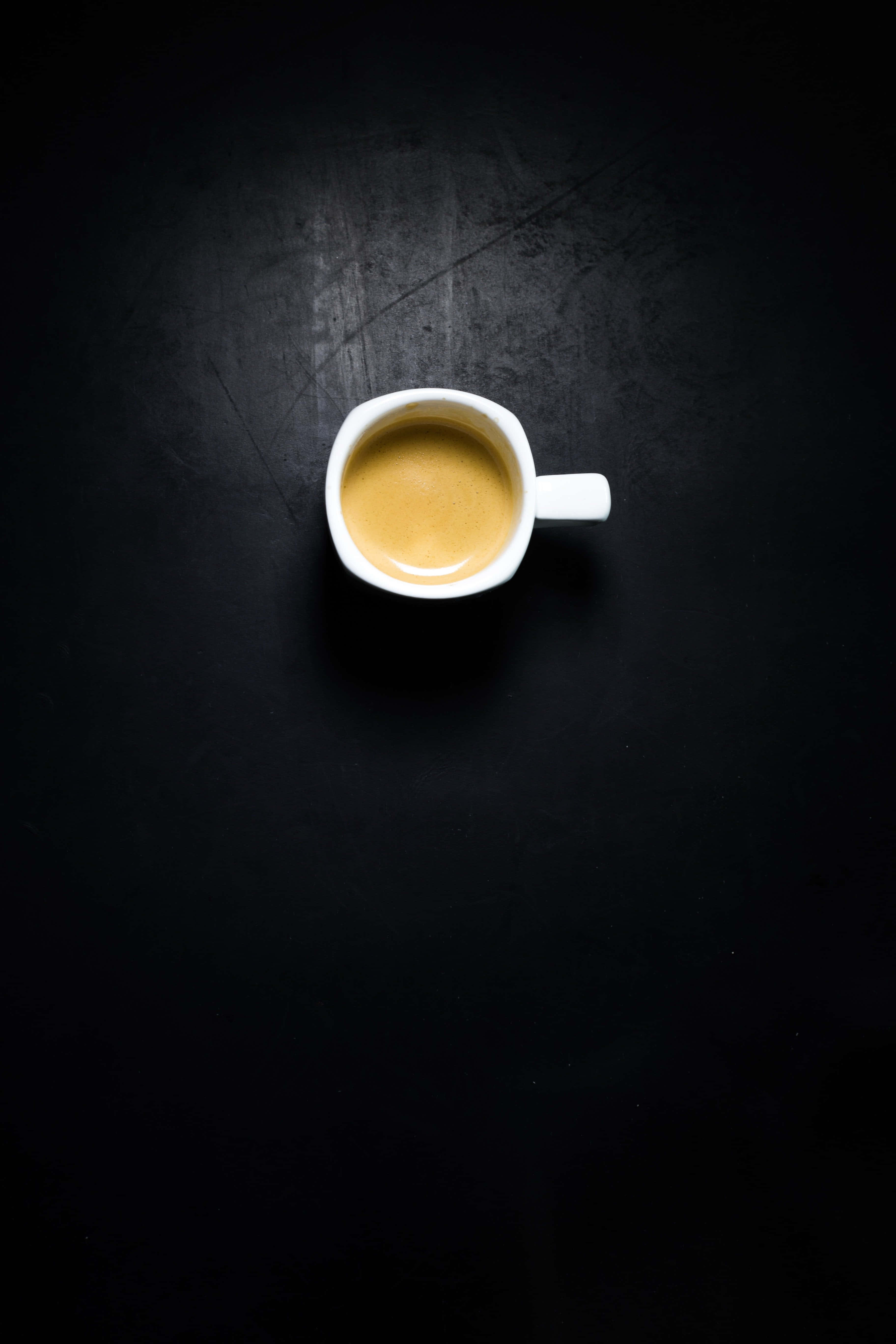 Black Coffee 桌布 壁紙 — 一個裝滿金色咖啡的白色濃縮咖啡杯，居中放置在帶有紋理的黑色表面上，並處於戲劇性的聚光燈下。