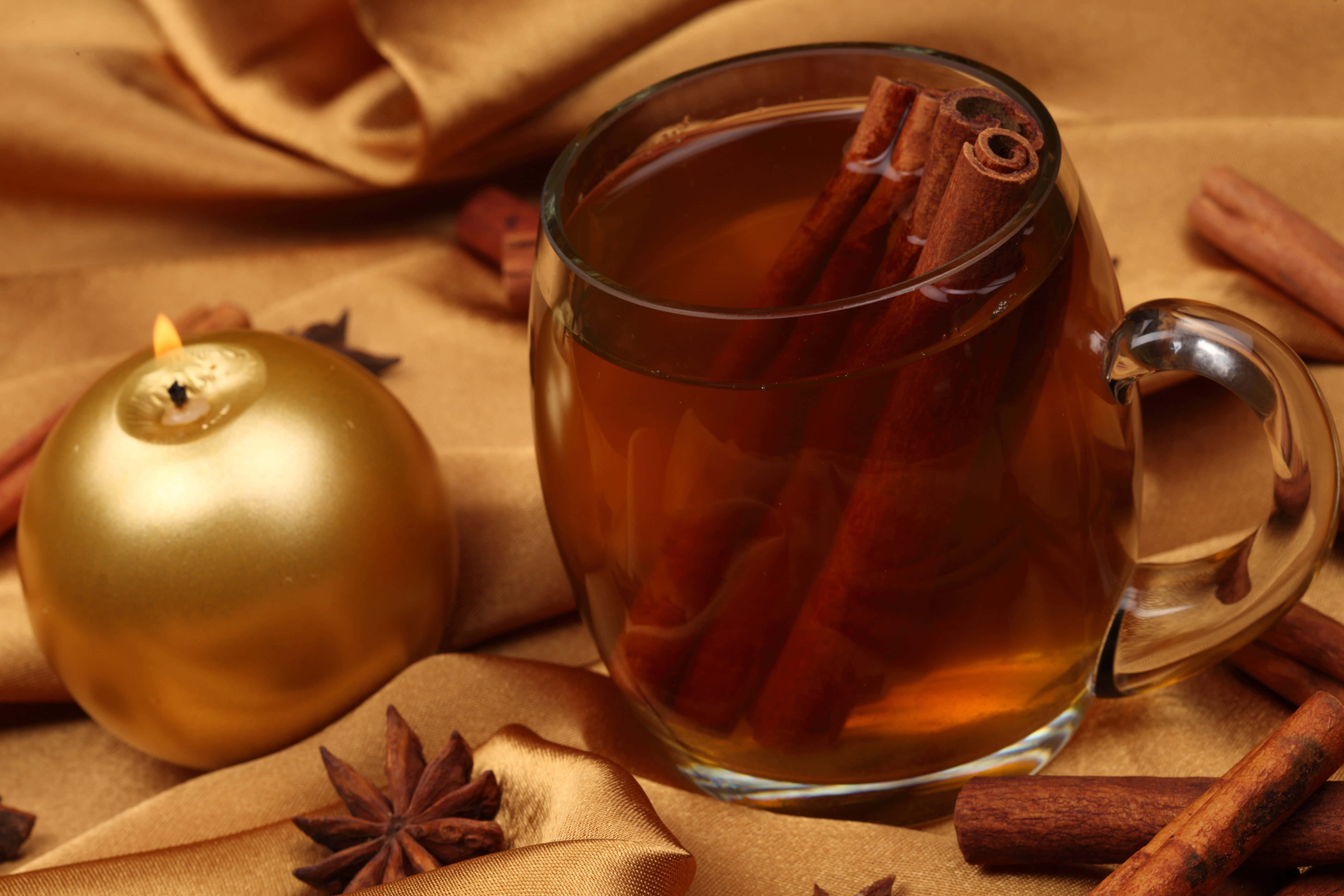 Mulled Wine 桌布 壁紙 — 一個裝滿香料茶和肉桂棒的溫暖玻璃杯，旁邊放著一支點燃的金色蠟燭，放在金色布料上。