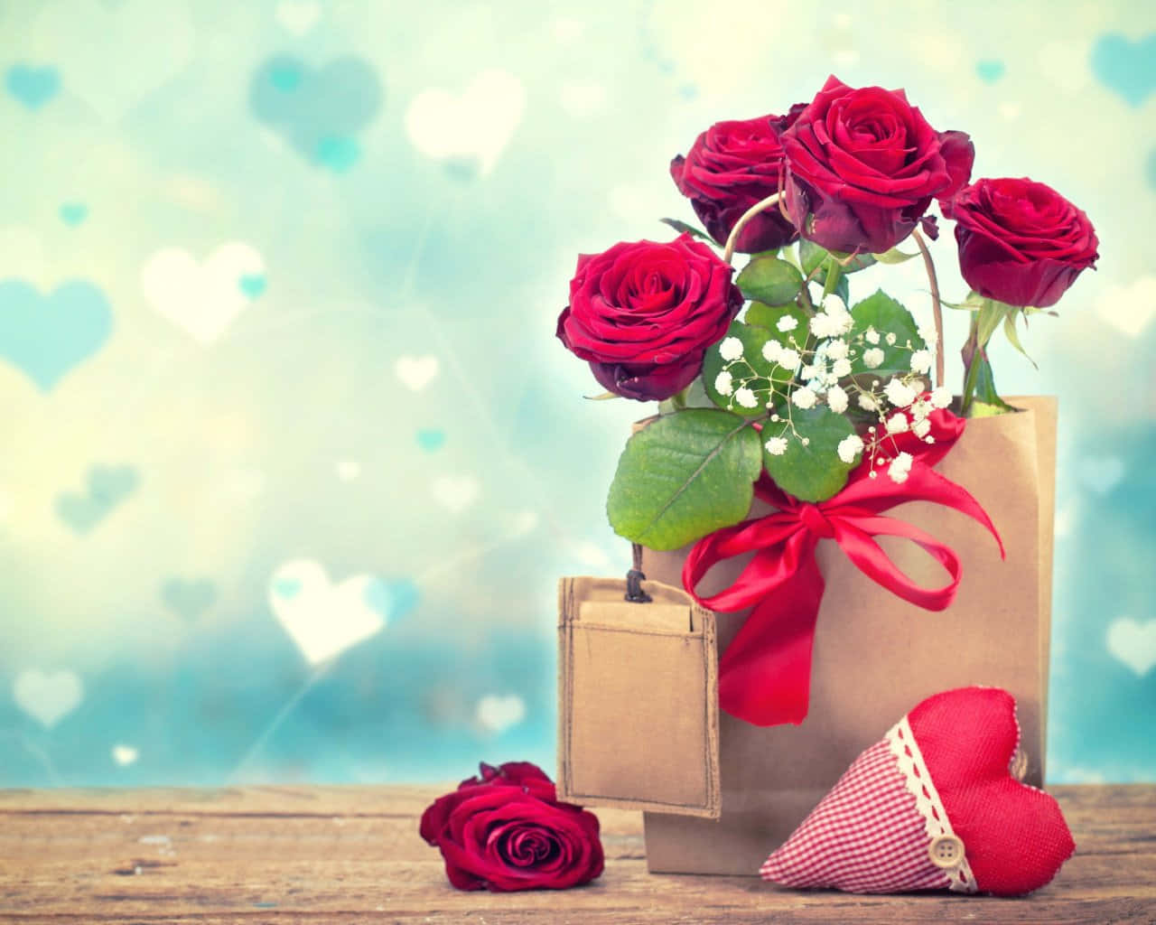 Rose Heart 桌布 壁紙 — 一個裝滿鮮豔紅玫瑰和滿天星的棕色紙袋放在木質表面上，旁邊有一個心形靠墊和一朵單獨的玫瑰，背景是柔和的藍色心形圖案。