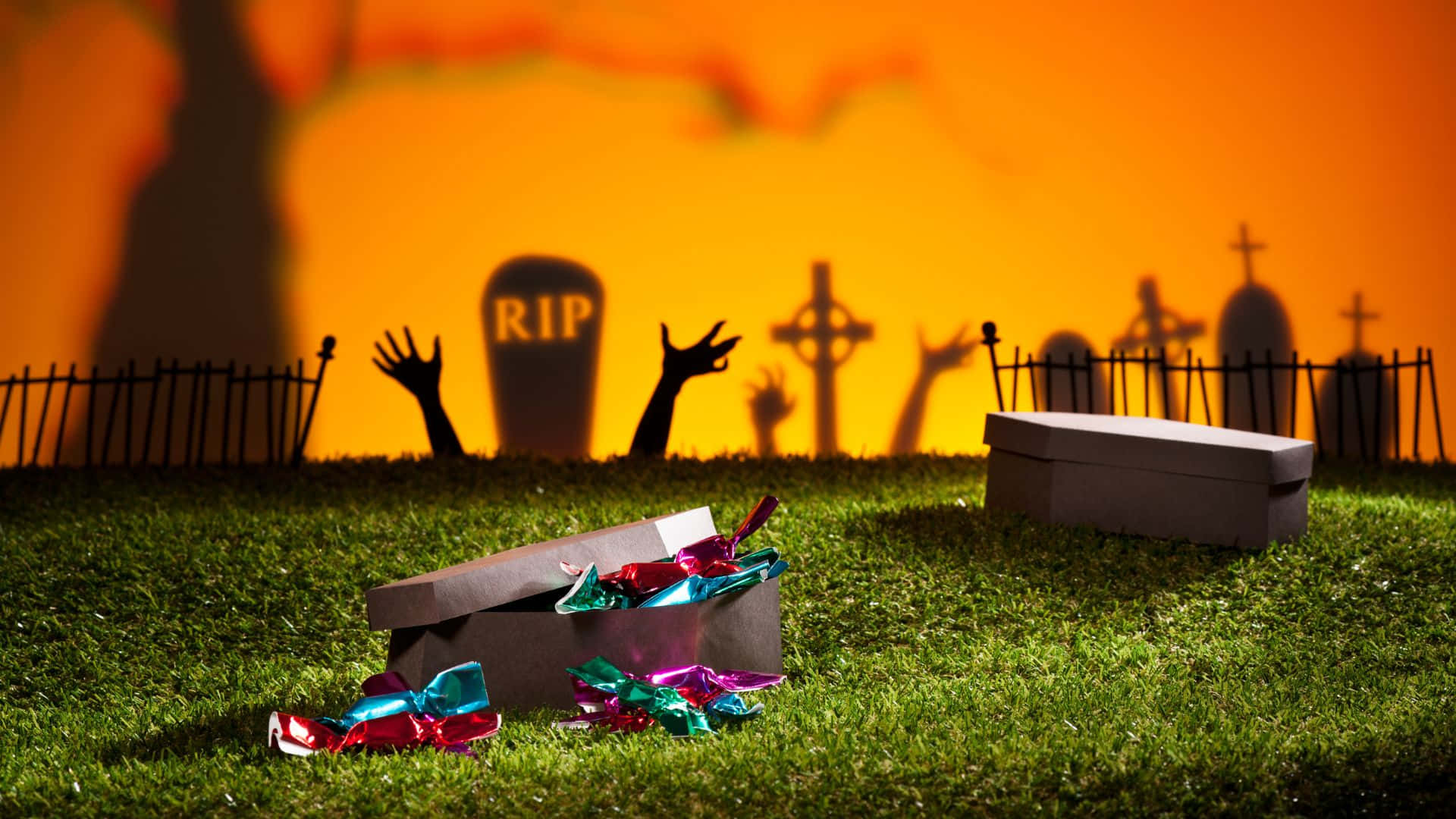 Halloween Coffins 桌布 壁紙 — 一個詭異的萬聖節墓地，有墓碑和殭屍手從草叢中升起，糖果從打開的棺材中灑出。