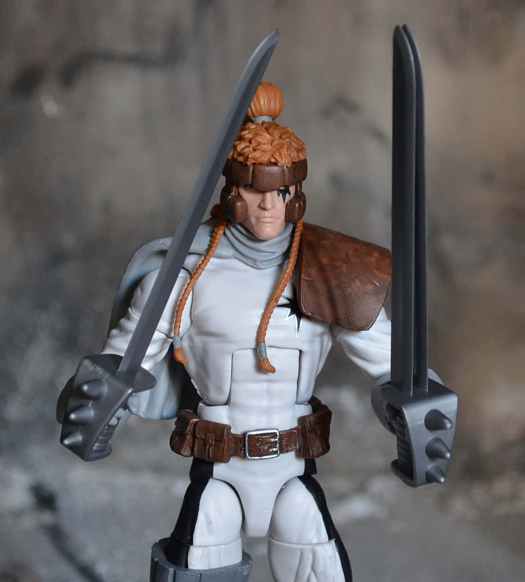 Shatterstar 桌布 壁紙 — 一個詳細嘅動作人偶，係一個有橙色辮子頭髮同兩把劍嘅戰士，企喺模糊嘅石頭背景前面。