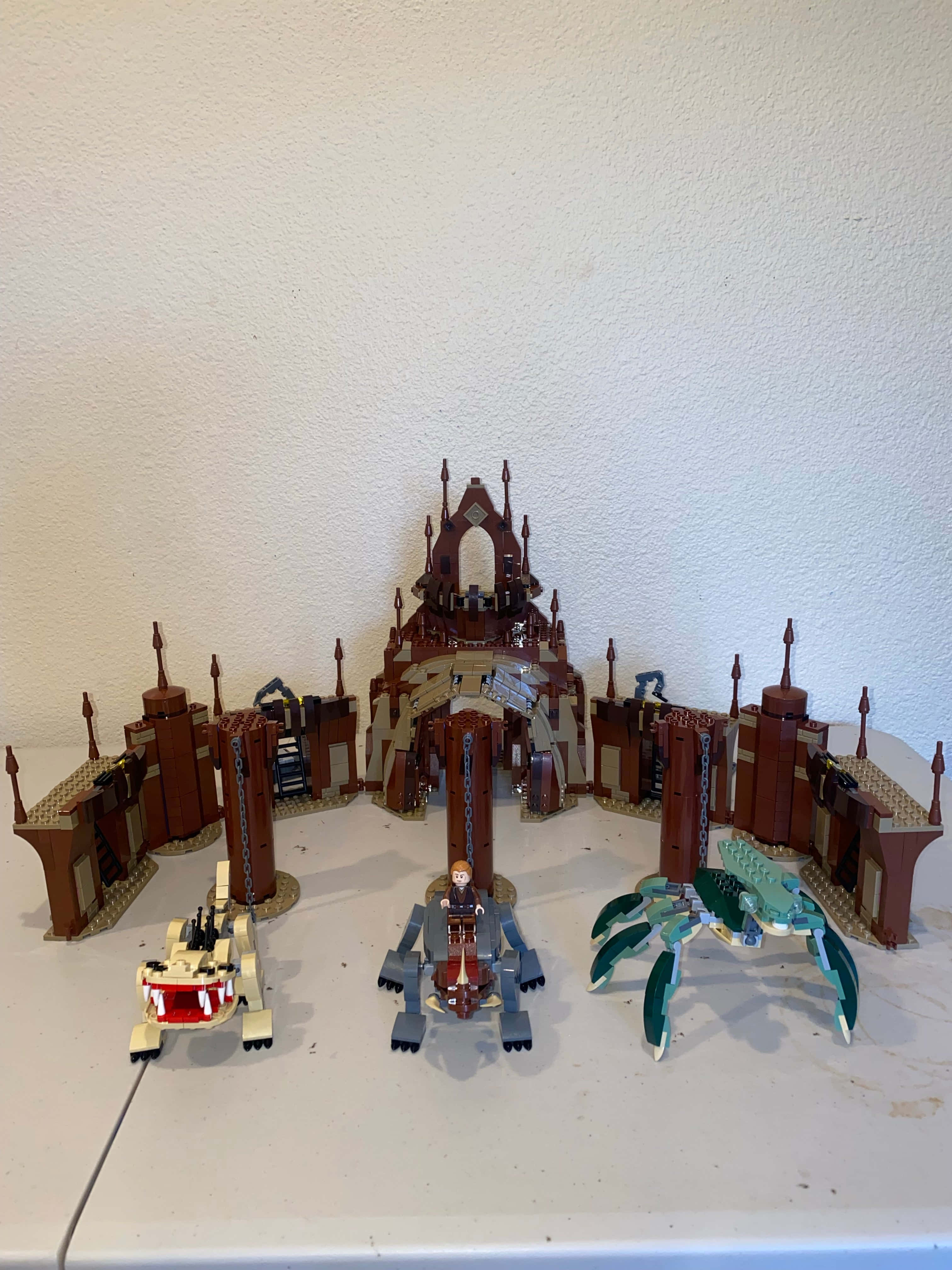 The Geonosian Arena 桌布 壁紙 — 一個詳細的LEGO Star Wars堡壘，三個車輛展示在白色桌子上，背景為有紋理的牆壁。