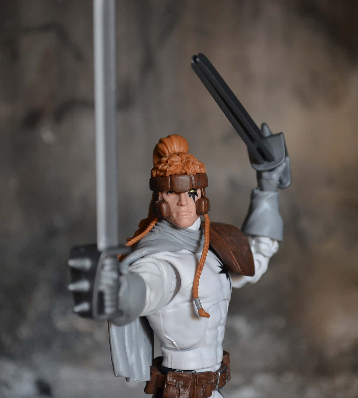 Shatterstar 桌布 壁紙 — 一個詳細的動作人偶，戰士有橙色辮子頭髮和頭帶，在模糊的岩石背景下揮舞劍和手槍。