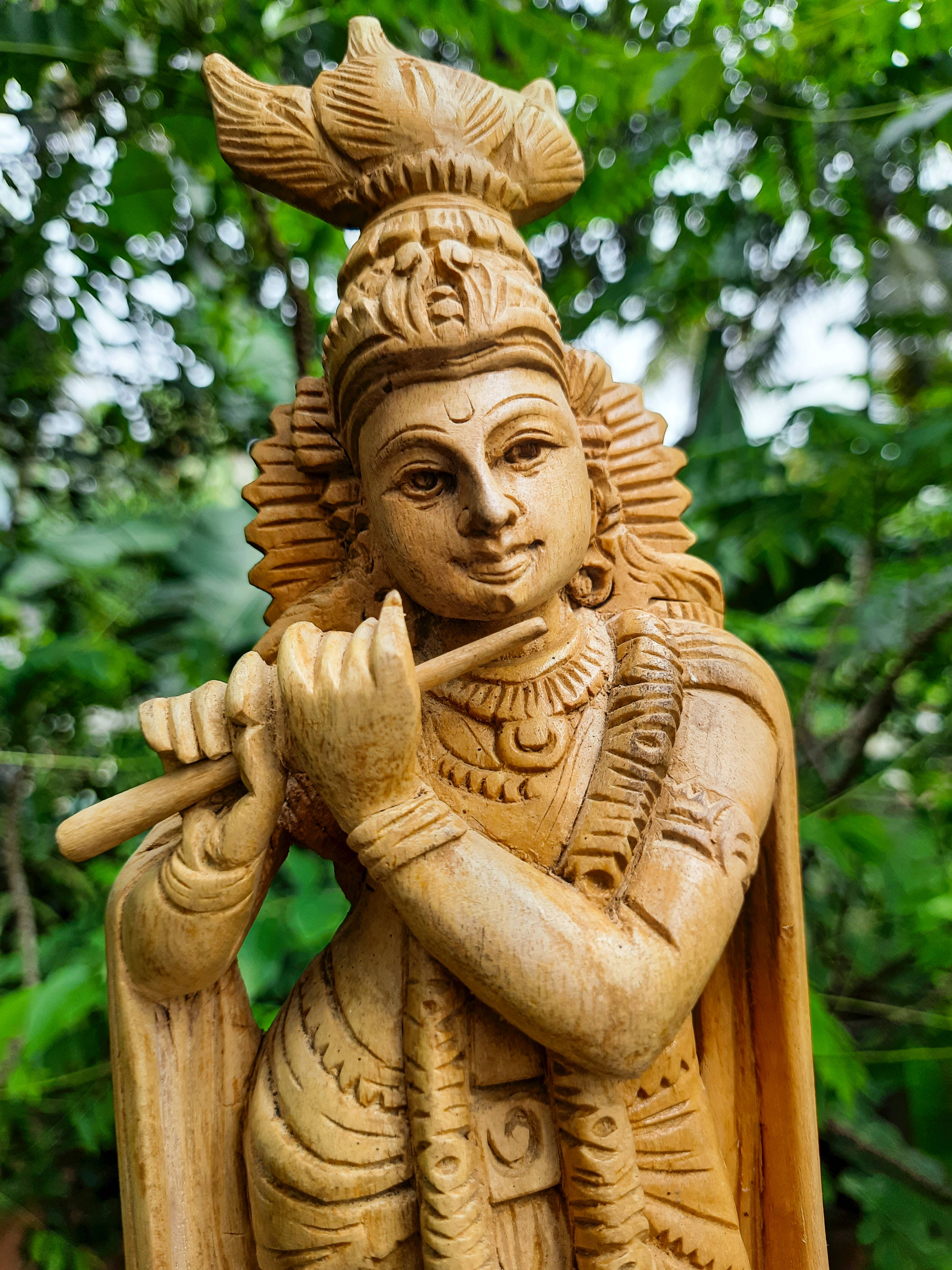 krishna iPhone 桌布 壁紙 — 一個詳細的木製雕像，描繪一位寧靜的神明吹奏長笛，矗立在茂密的綠色樹葉中。