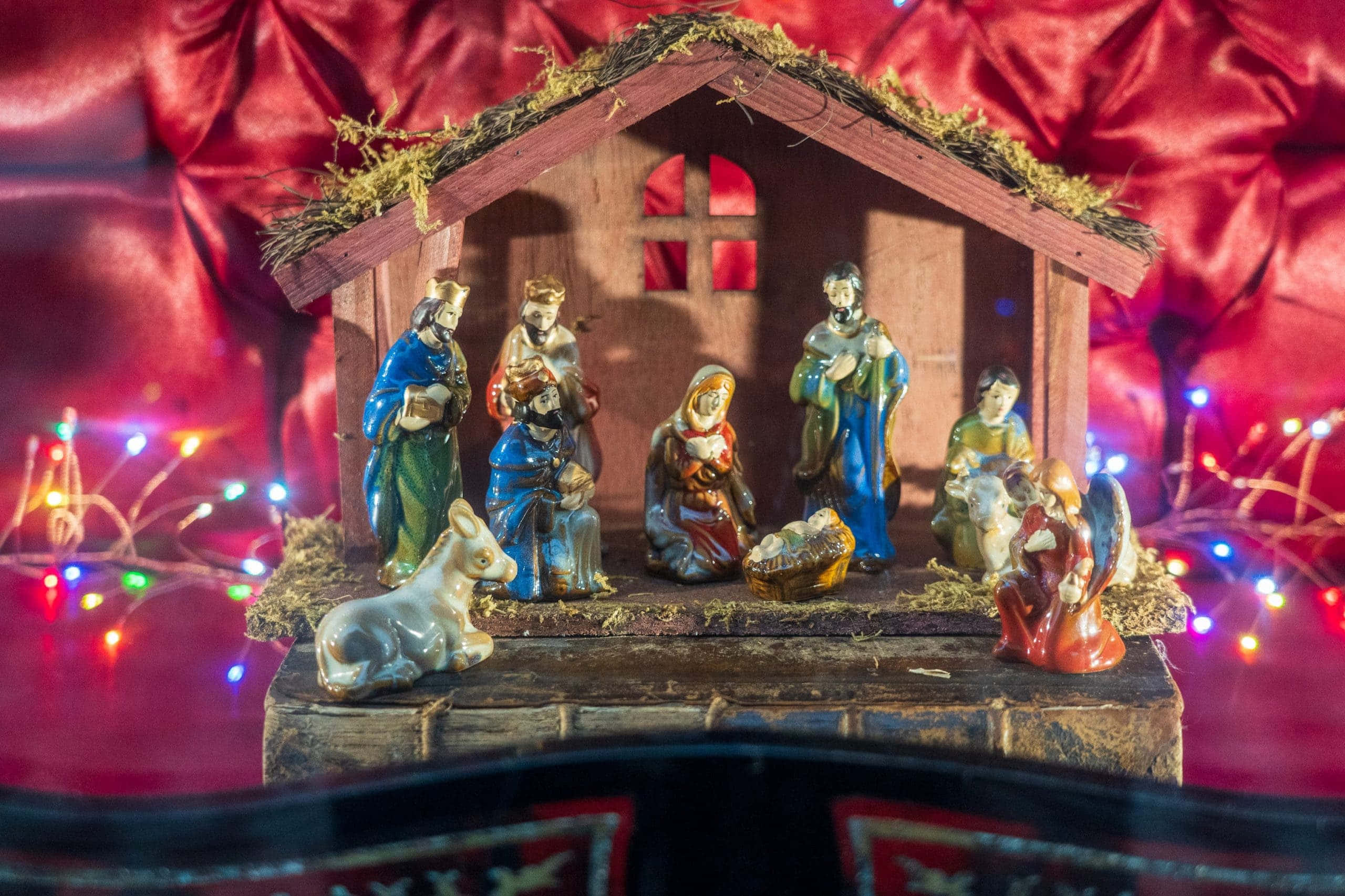 Nativity Scene 桌布 壁紙 — 一個詳細的耶穌誕生場景，人物在木製馬廄屋頂下，周圍是閃爍的仙女燈，背景是濃郁的紅色緞面。