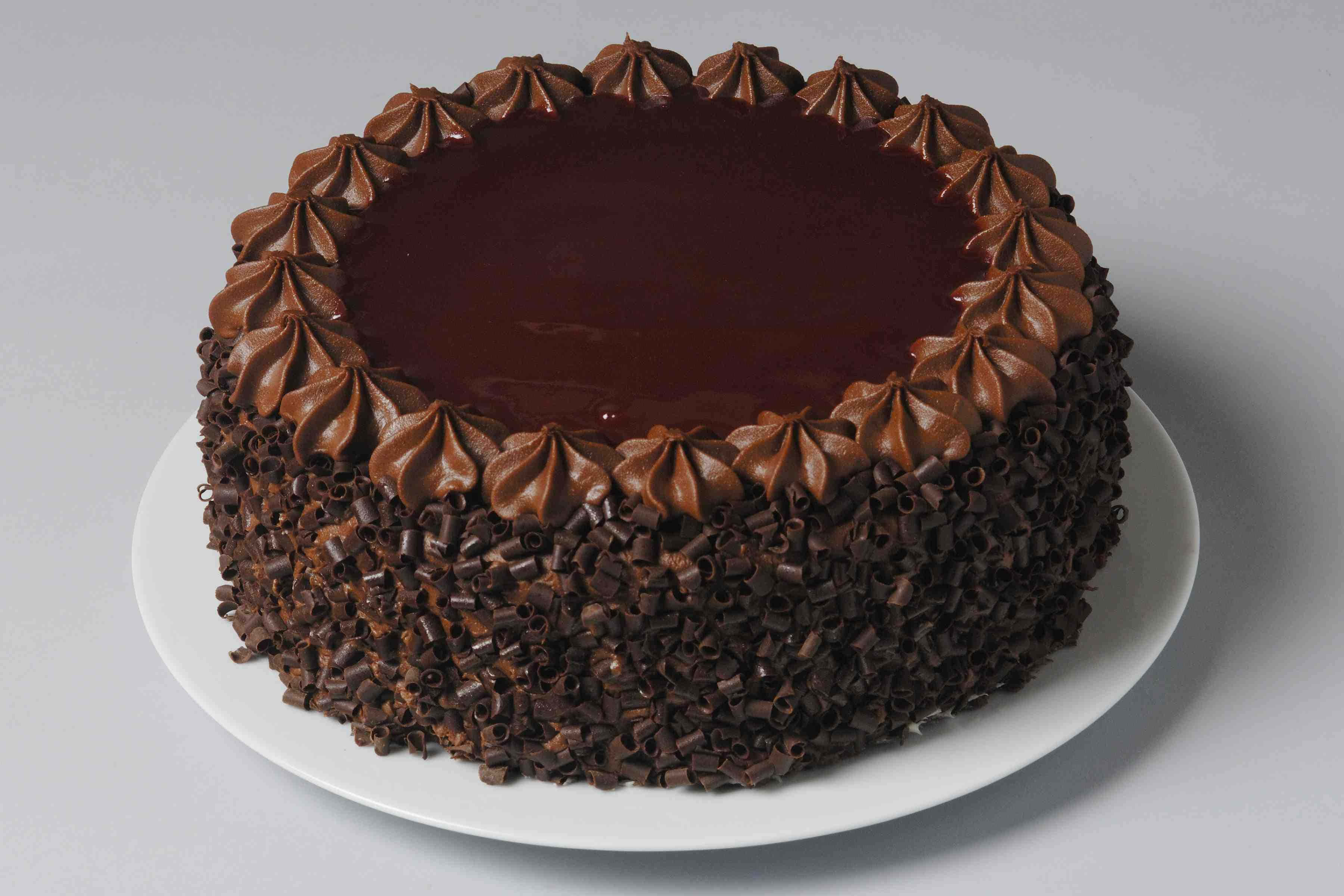 Chocolate Cake 桌布 壁紙 — 一個豐富的朱古力蛋糕，帶有光澤的甘納許頂部、擠花玫瑰和朱古力卷，放在白色碟子上。