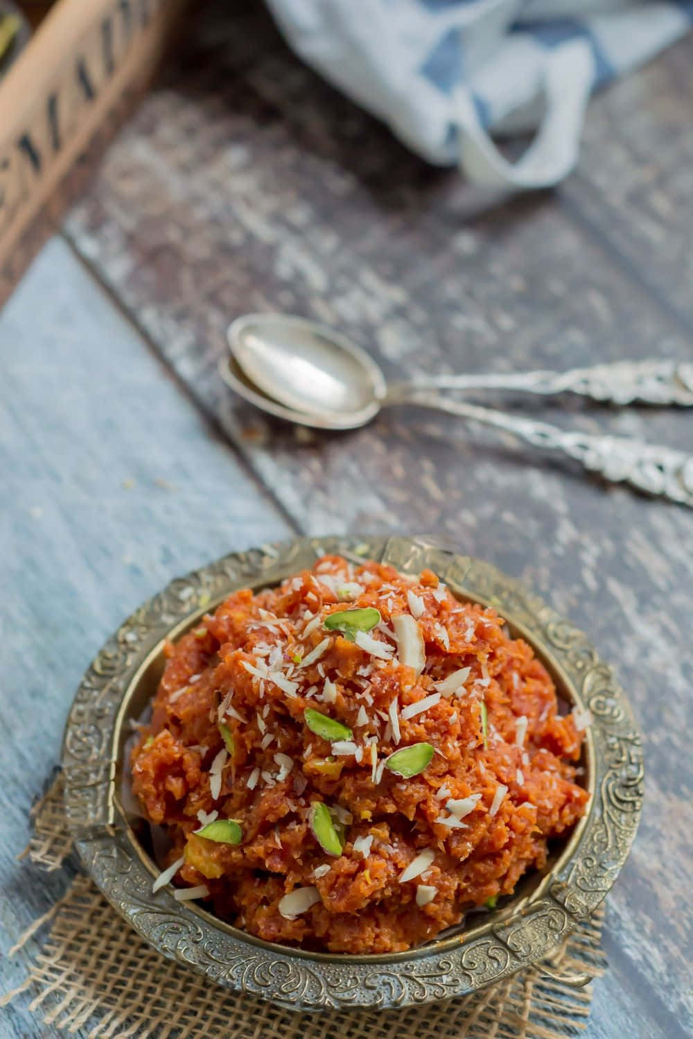 Indian Food 桌布 壁紙 — 一個豐富的橙色紋理菜餚，上面放有切片杏仁和開心果，放在一個裝飾性的金屬碗中，置於鄉村風格的木質表面上。