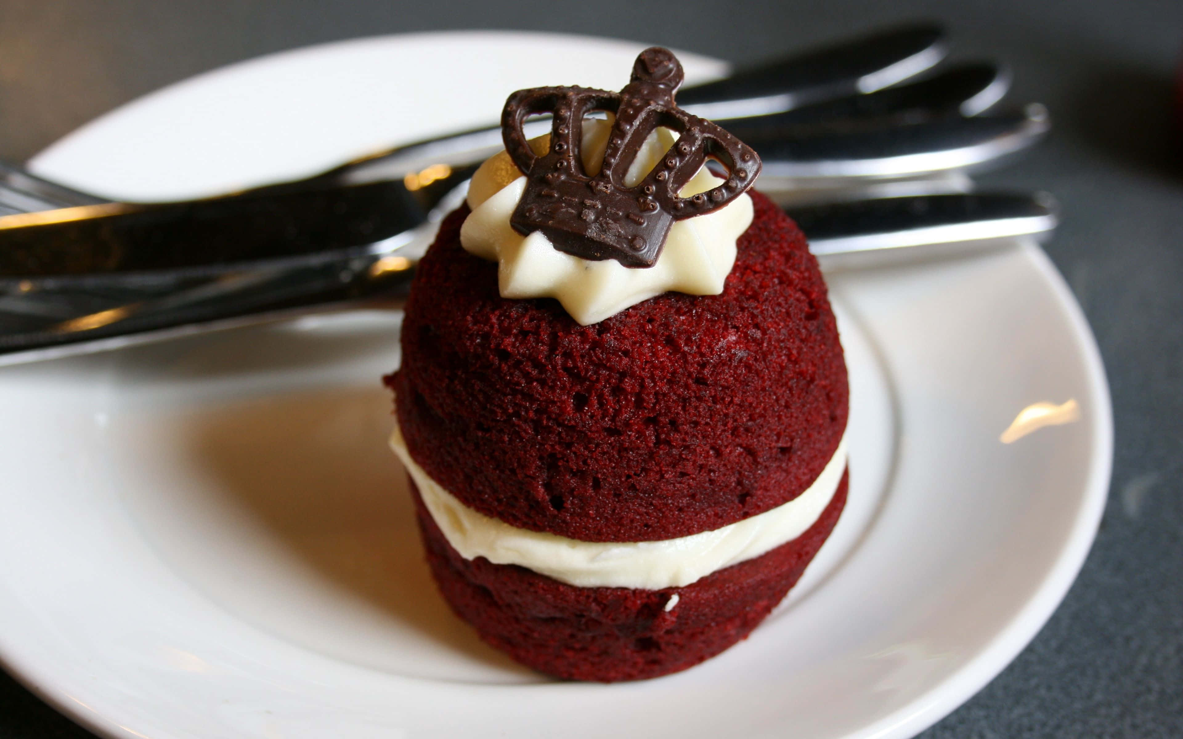 Red Velvet Cake 桌布 壁紙 — 一個豐富的紅絲絨杯子蛋糕，內有白色奶油餡料和朱古力皇冠，優雅地放在一個白色碟子上，旁邊有銀叉。