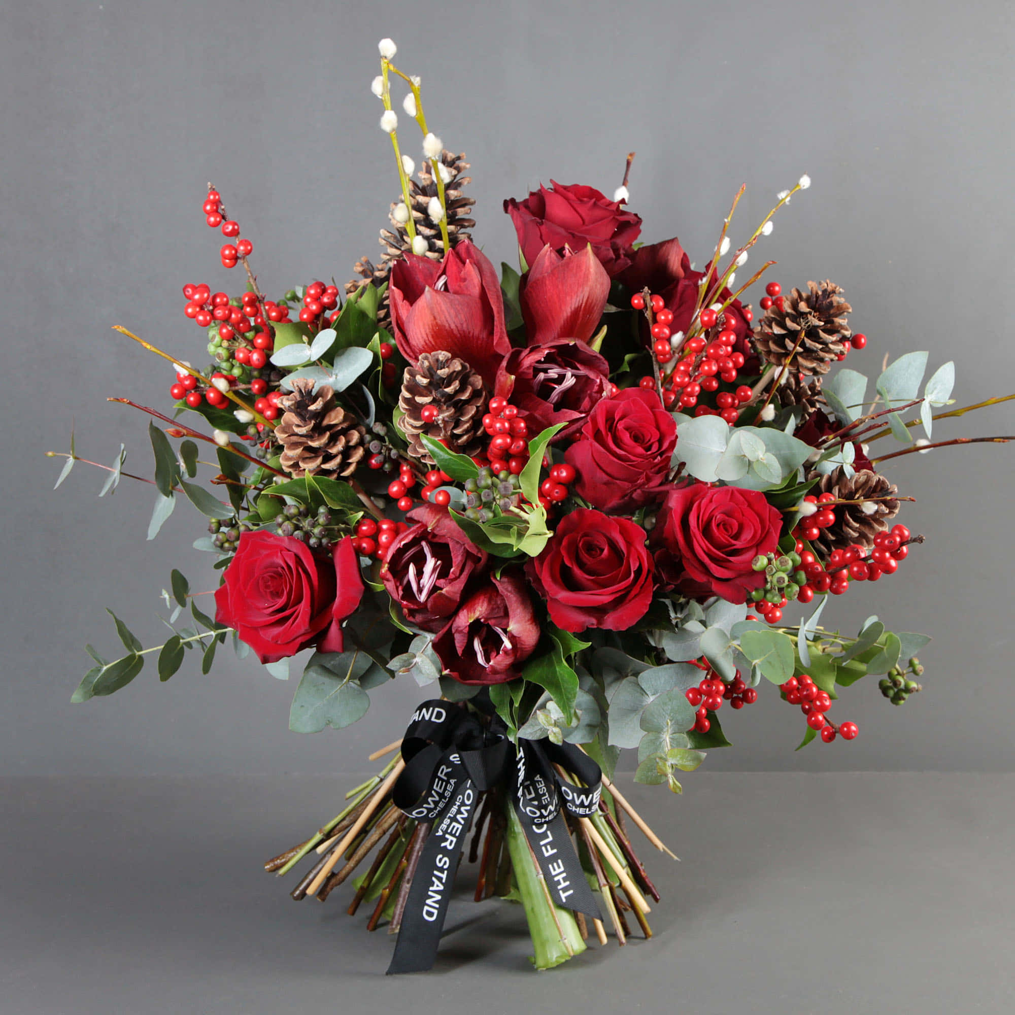 Flower Bouquet 桌布 壁紙 — 一個豐盛的節日花束，包含深紅玫瑰和鬱金香，點綴著松果和鮮紅漿果，背景為中性灰色。