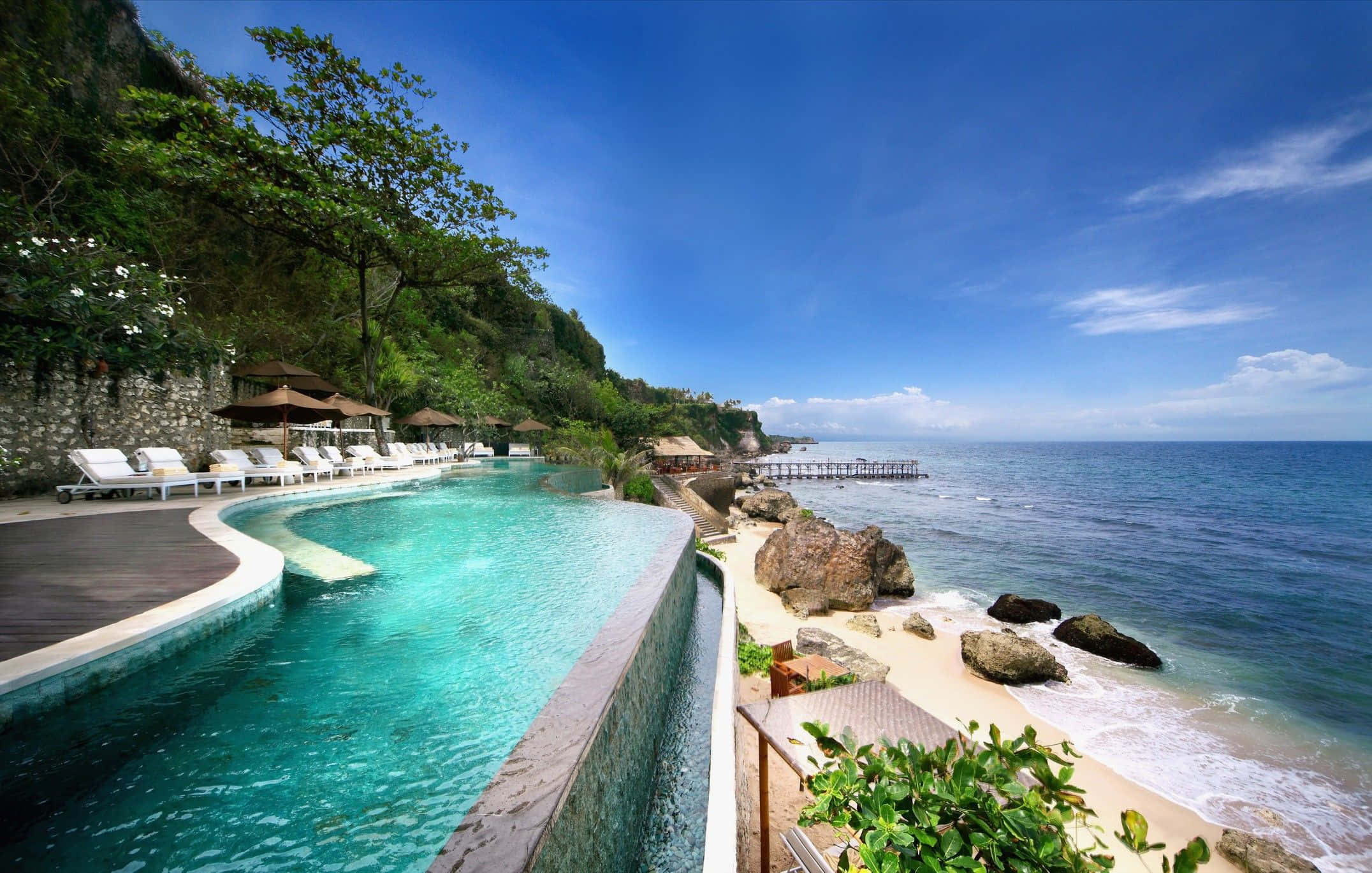 Bali Beach 桌布 壁紙 — 一個豪華的無邊際泳池，在清澈的藍天下，沿著岩石海灘流入碧綠的海洋。