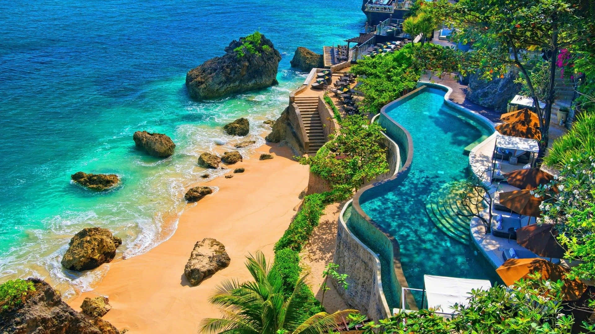 Bali Beach 桌布 壁紙 — 一個豪華的無邊際泳池沿著熱帶懸崖蜿蜒，俯瞰著碧綠的海水和散落著岩石的沙灘。