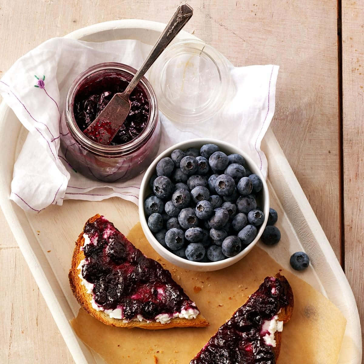 Blueberry Jam 桌布 壁紙 — 一個質樸的托盤上放著新鮮藍莓、一瓶自製藍莓果醬和塗上忌廉及果醬的多士片，置於木桌上。