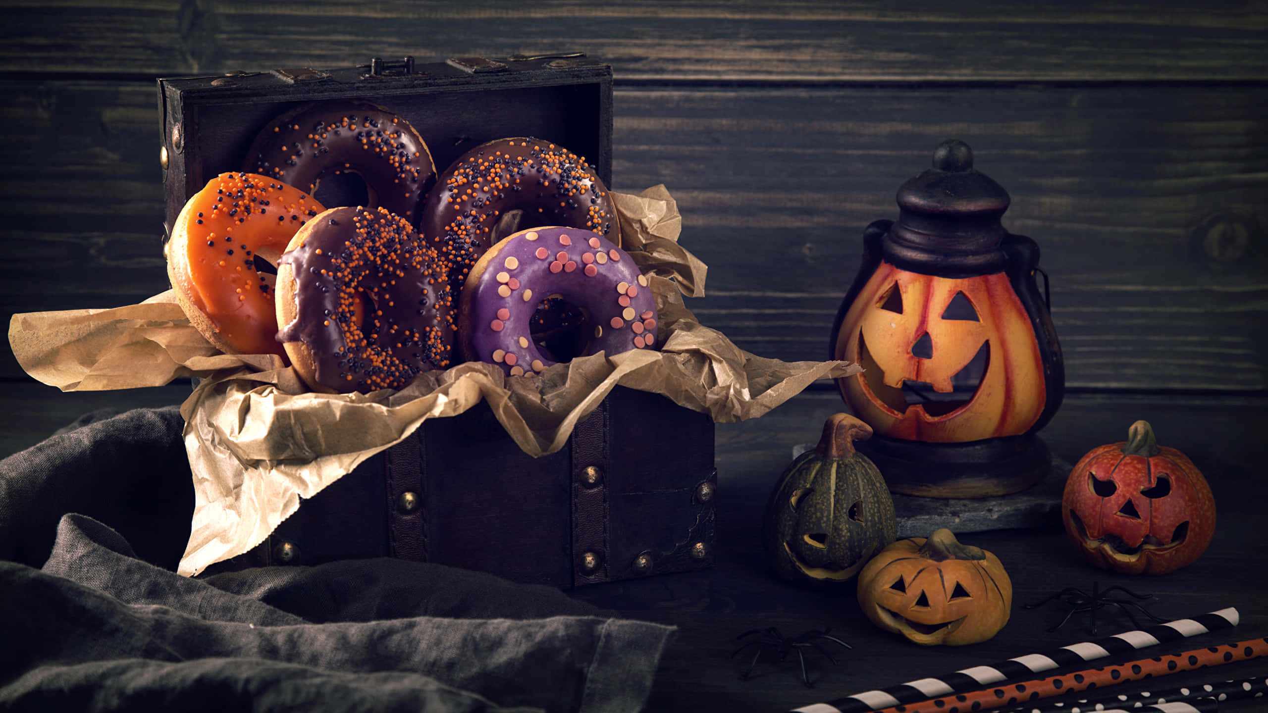 Halloween Treats 桌布 壁紙 — 一個質樸的木箱裝滿色彩繽紛的萬聖節冬甩，旁邊有一個發光的傑克南瓜燈和雕刻南瓜，放在深色木質表面上。
