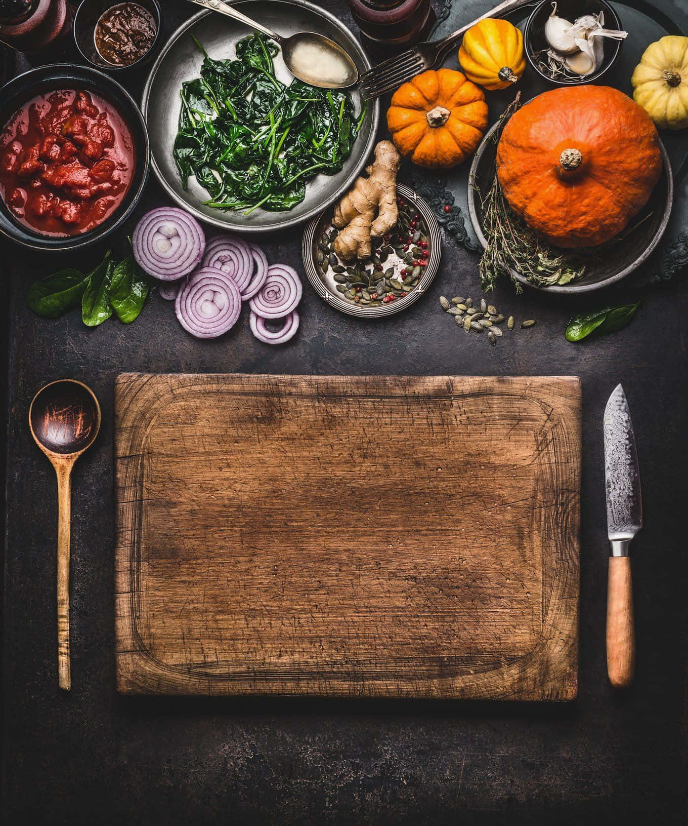 Winter Food 桌布 壁紙 — 一個質樸的木製砧板，置於南瓜、洋蔥和香料等秋季食材中央，放在深色紋理表面上。