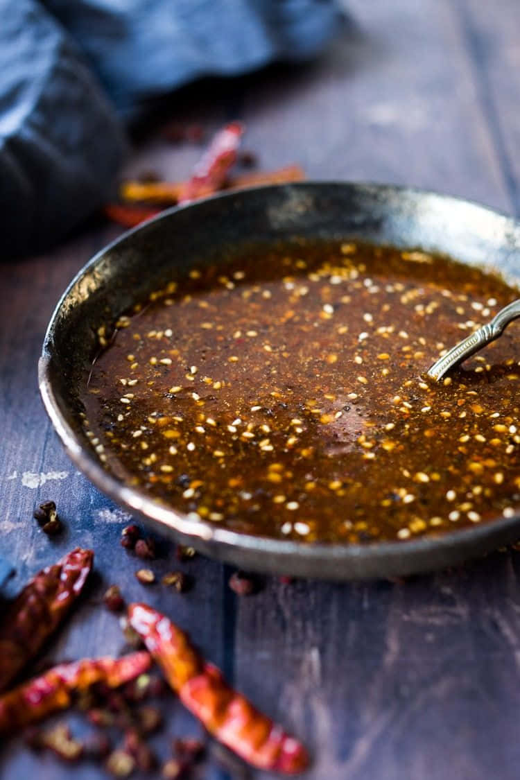Szechuan Sauce 桌布 壁紙 — 一個質樸的碗裡盛著濃郁的深色醬汁，上面撒滿芝麻和乾辣椒，放在風化的木質表面上。