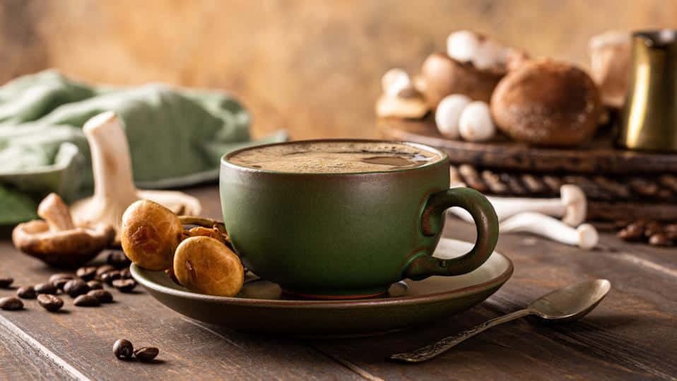Brown Coffee 桌布 壁紙 — 一個質樸的綠色陶瓷咖啡杯放在木桌上，周圍有蘑菇、咖啡豆和復古勺子。