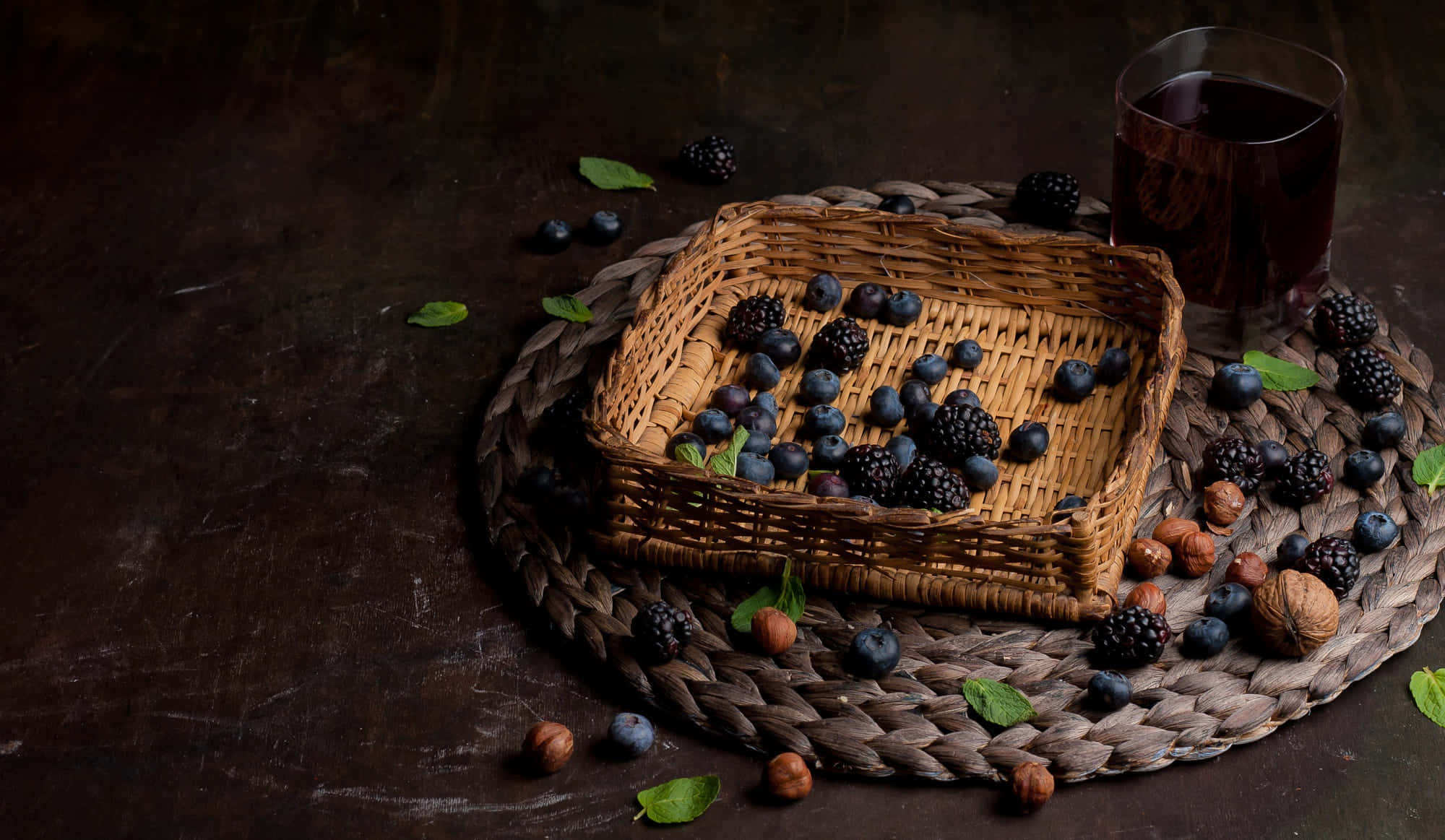 Blackberry Wine 桌布 壁紙 — 一個質樸的編織籃子裝滿了黑莓和藍莓，旁邊放著一杯深色果汁，放在深色紋理表面上。