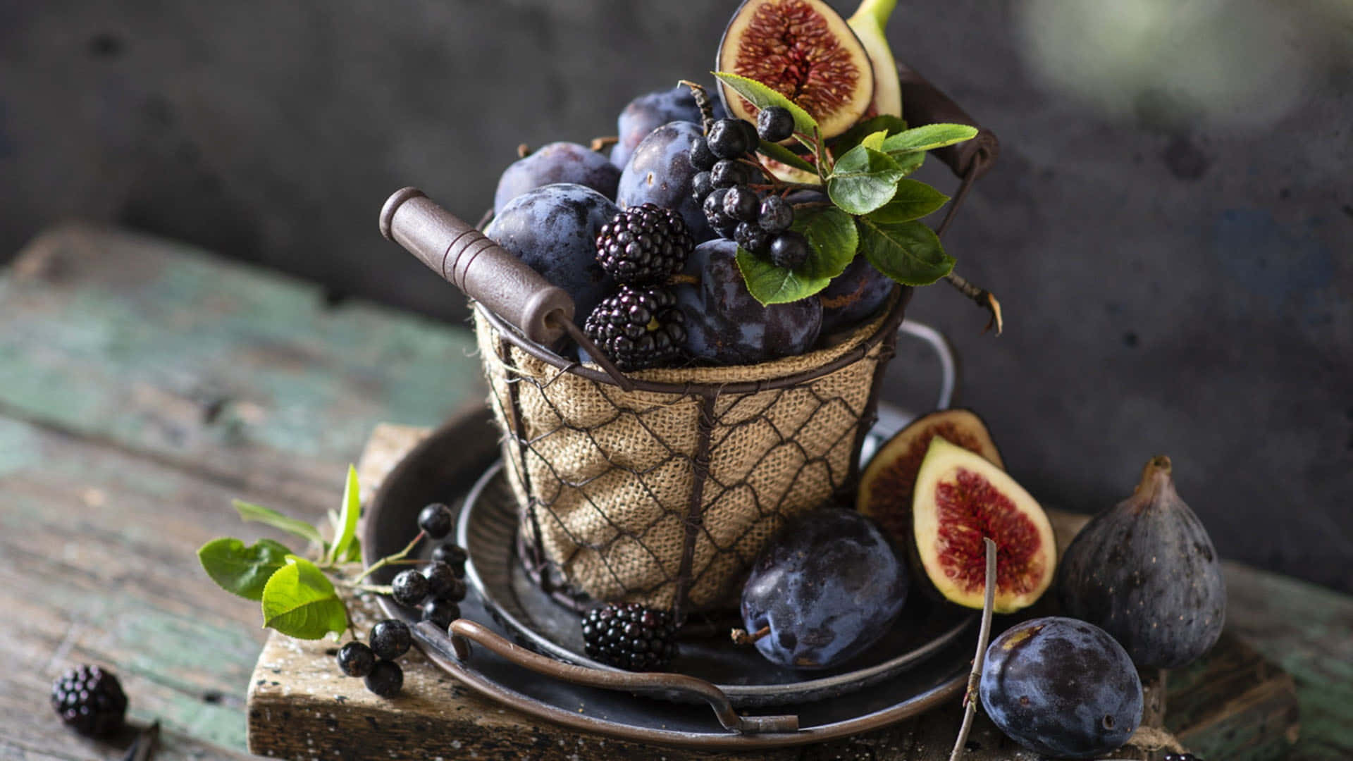 Purple Figs 桌布 壁紙 — 一個質樸的鐵絲籃子裝滿了李子、黑莓和無花果，放在一張風化的木桌上。