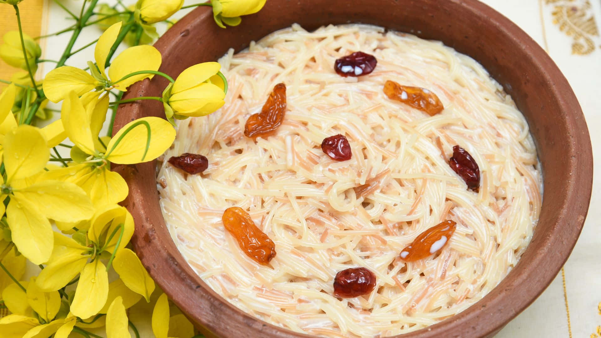 Vishu 桌布 壁紙 — 一個質樸的陶碗盛載著奶油狀的碎甜點，上面放有金色和深色葡萄乾，旁邊是鮮豔的黃色花朵。
