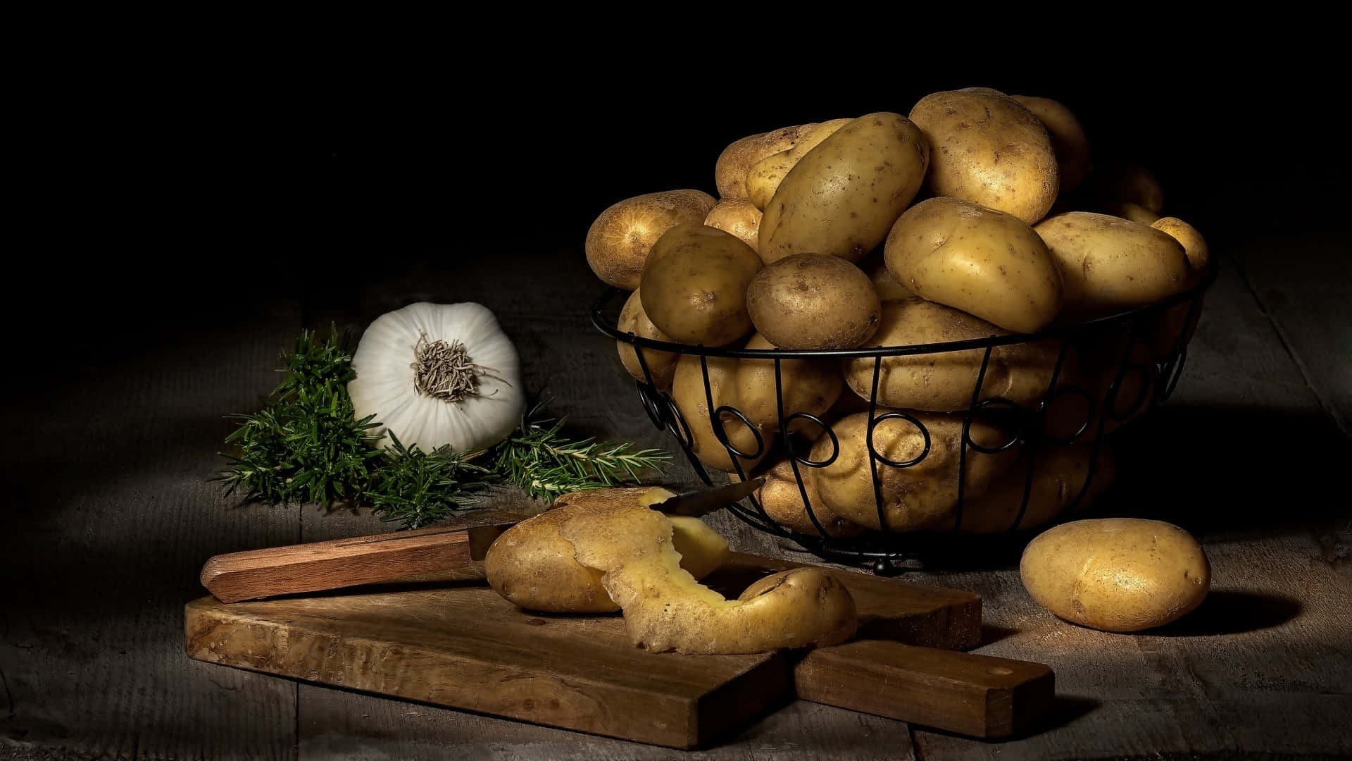 Potato 桌布 壁紙 — 一個質樸的靜物，馬鈴薯、大蒜和香草在深色木桌上沐浴在戲劇性的聚光燈下。