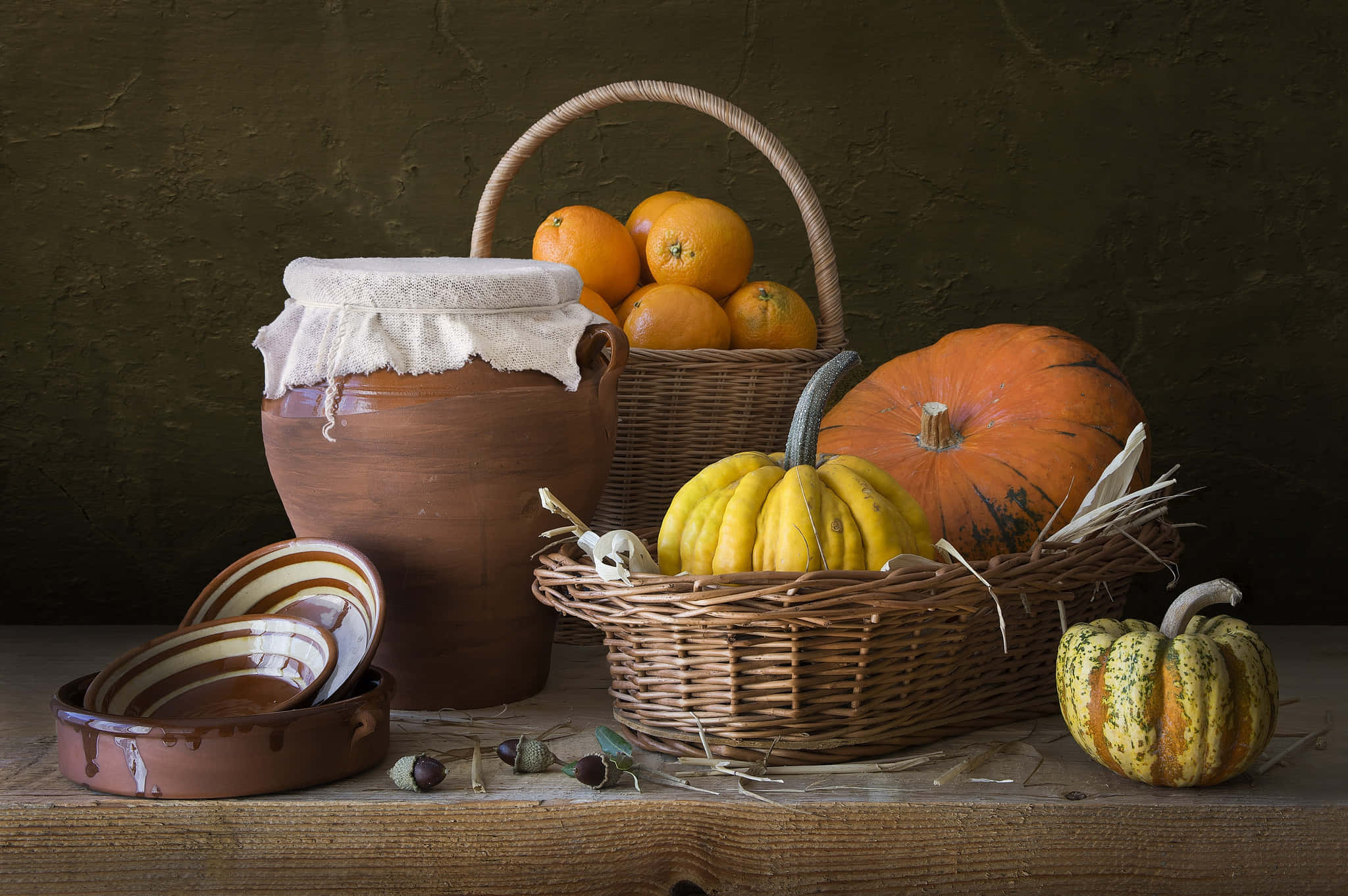 Fall Pumpkin 桌布 壁紙 — 一個質樸的靜物場景，南瓜、橙子和陶瓷碗擺放在木桌上，背景是深色有紋理的牆壁。
