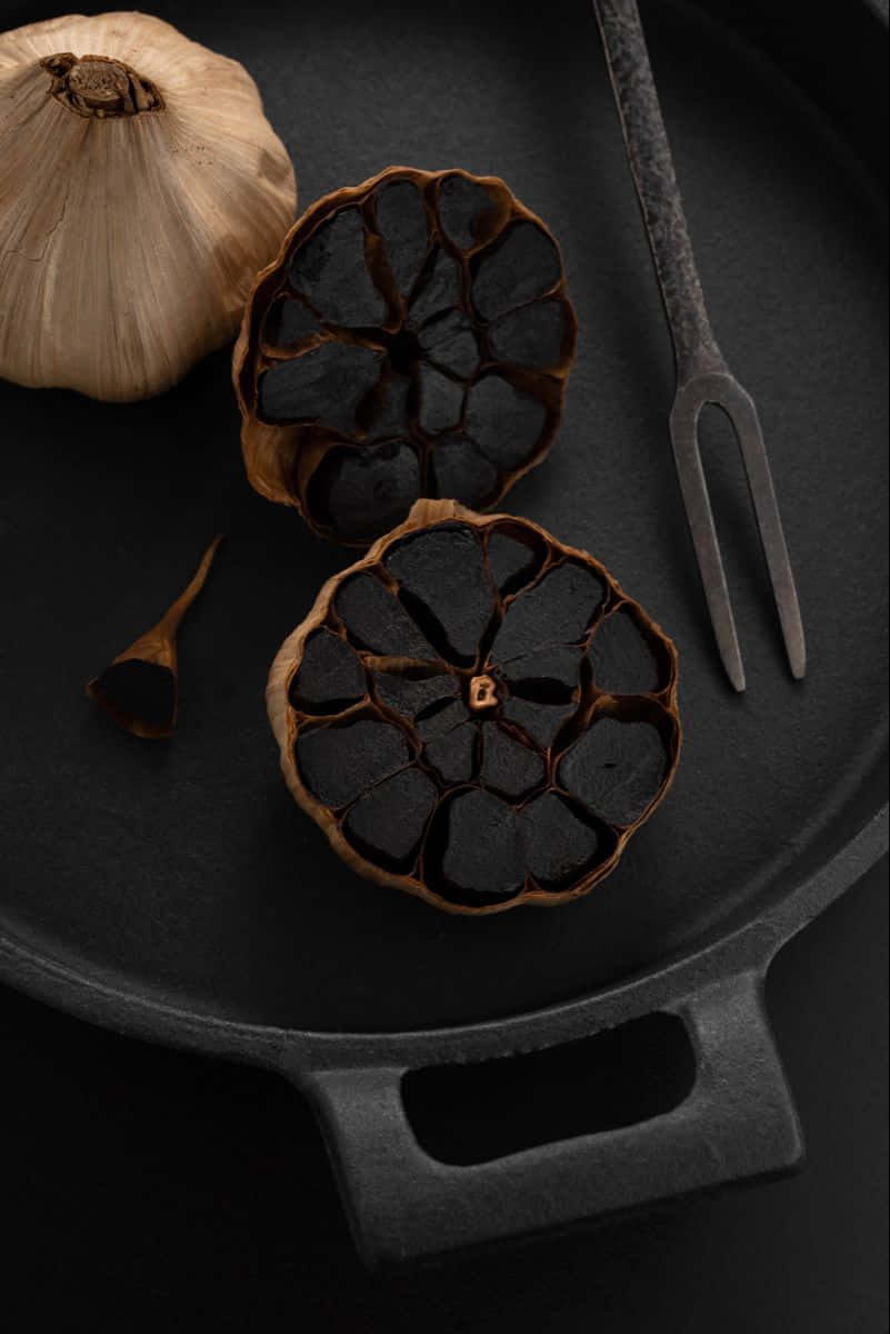 Black Garlic 桌布 壁紙 — 一個質樸的黑色鑄鐵煎鍋上放著烤大蒜球和一個復古叉子，背景為深色。