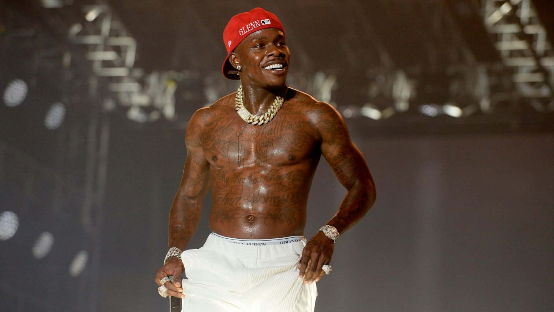 Dababy 桌布 壁紙 — 一個赤裸上身、有紋身、戴紅帽的男人在昏暗的舞台上咧嘴大笑，手持麥克風。